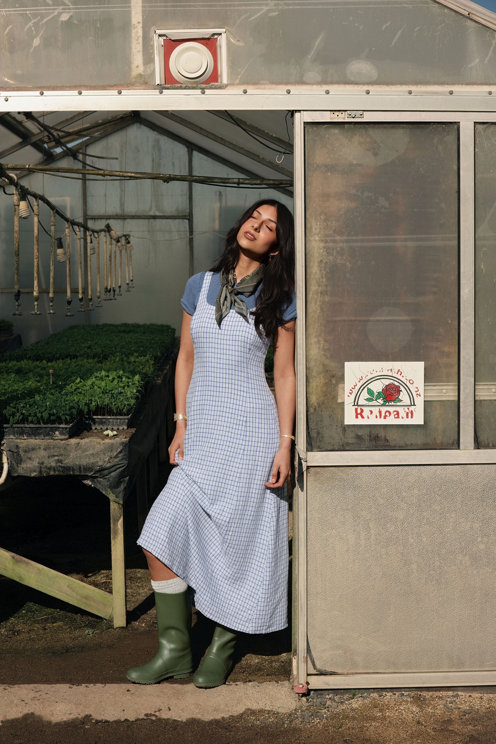 A woman standing in a greenhouse wearing a checkered spring midi dress.