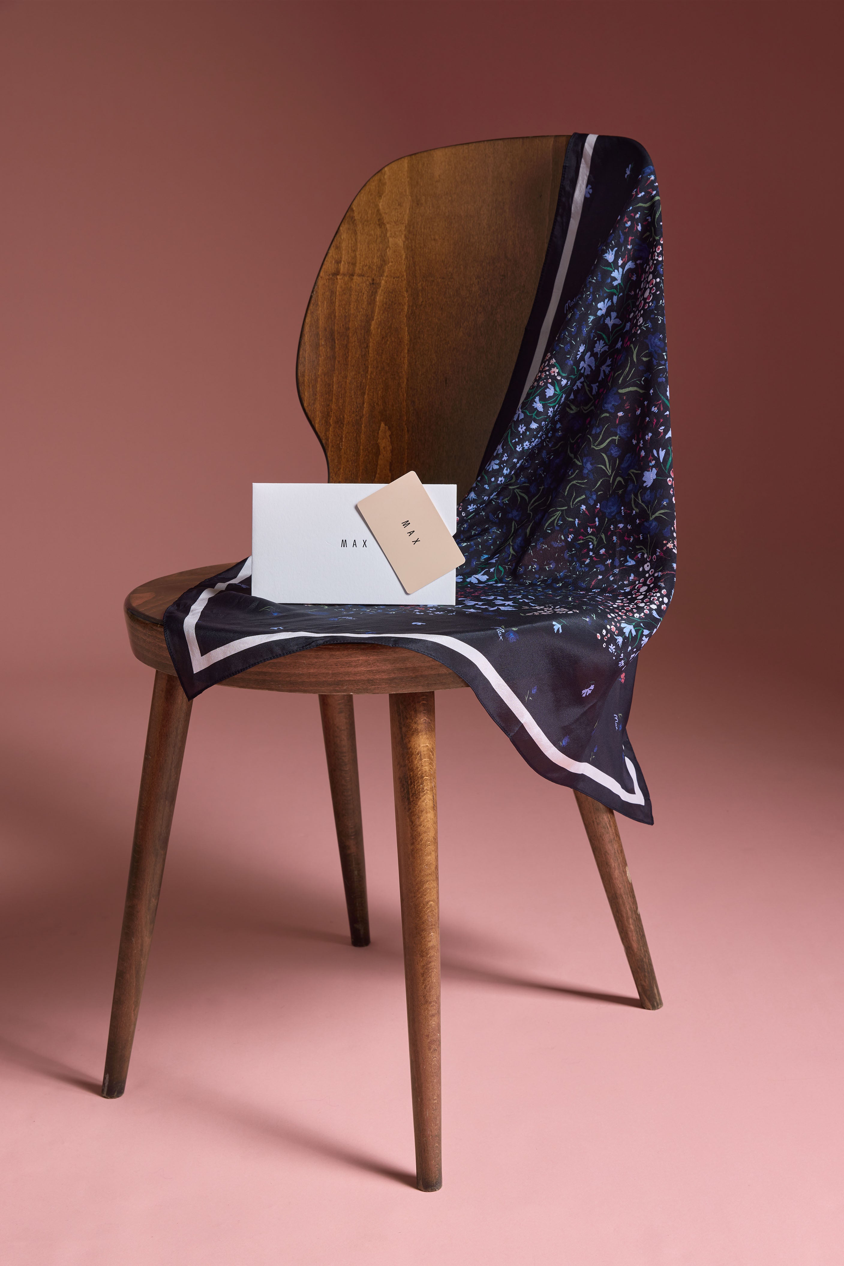 A wooden chair with a Max Printed Gift Card, dark floral scarf, and white box on pink.