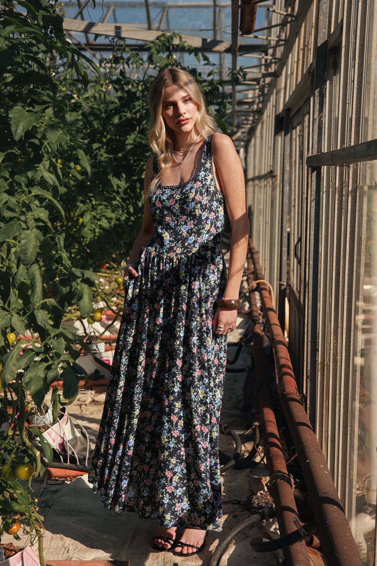Woman in Max’s Aria Gathered Waist Midi Dress stands in a sunny greenhouse with leafy plants.
