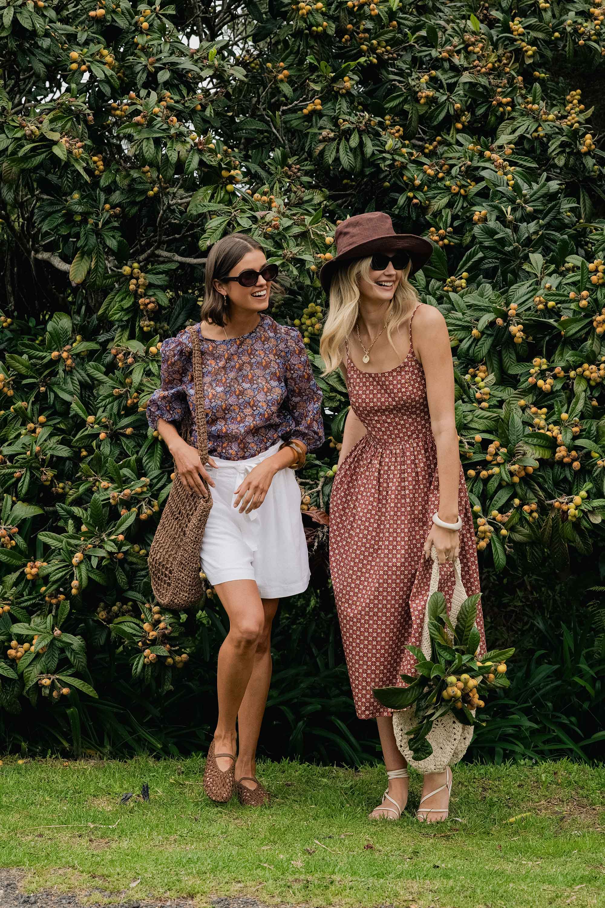 Two women in Max Eliza Midi Dresses and sunglasses smile by a leafy tree.