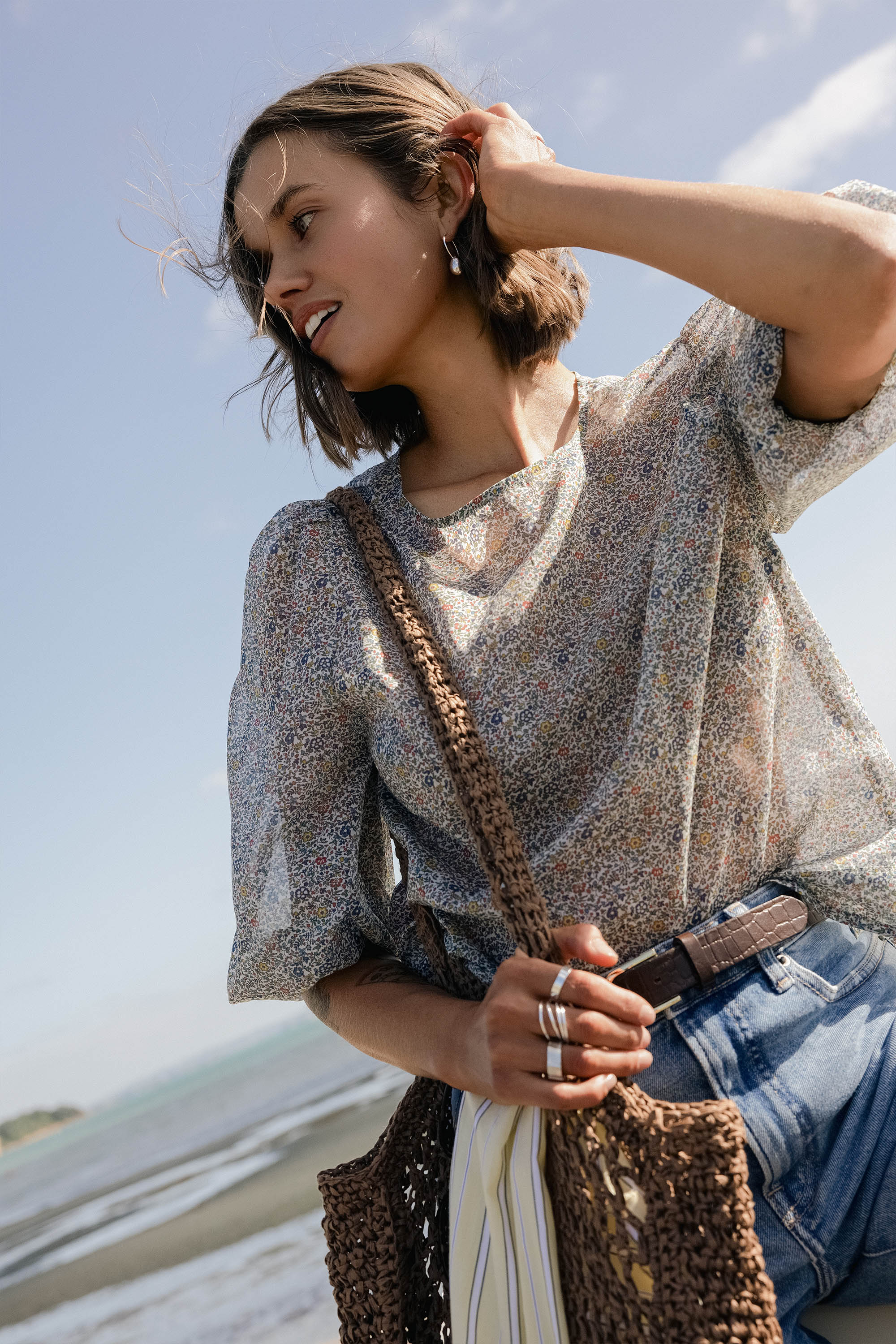 Woman in a Max Eloise Puff Sleeve Blouse and jeans stands on the beach with a woven bag.