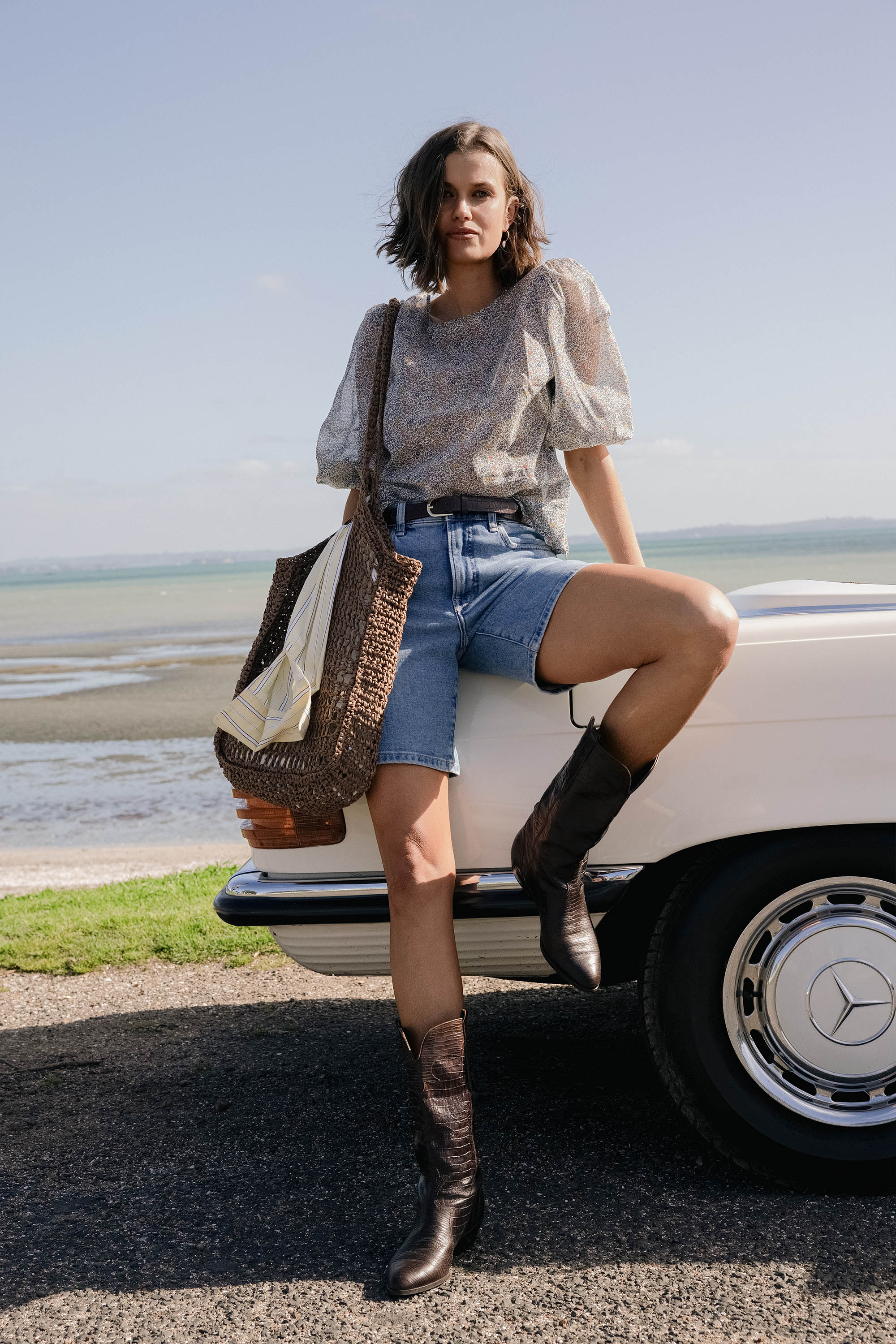Woman in Max Eloise Puff Sleeve Blouse, denim shorts, and boots leans on a white car by water.