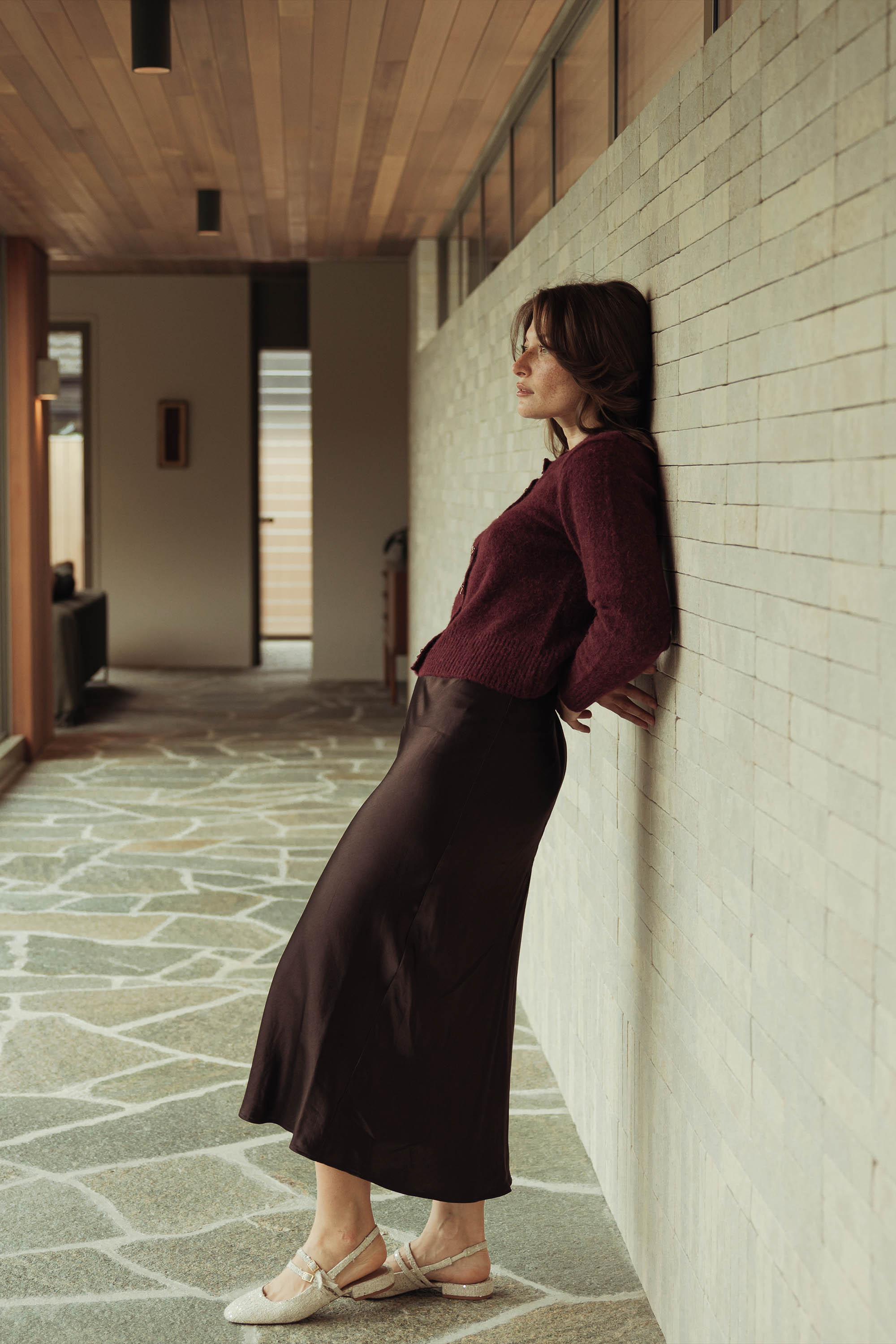 Woman in maroon top and Max The Slip Skirt leans against a hallway wall, looking thoughtful.