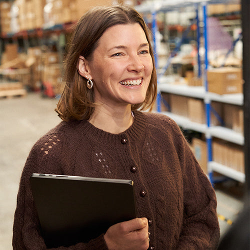 Smiling woman holding a laptop stands in a warehouse with shelves in the background.