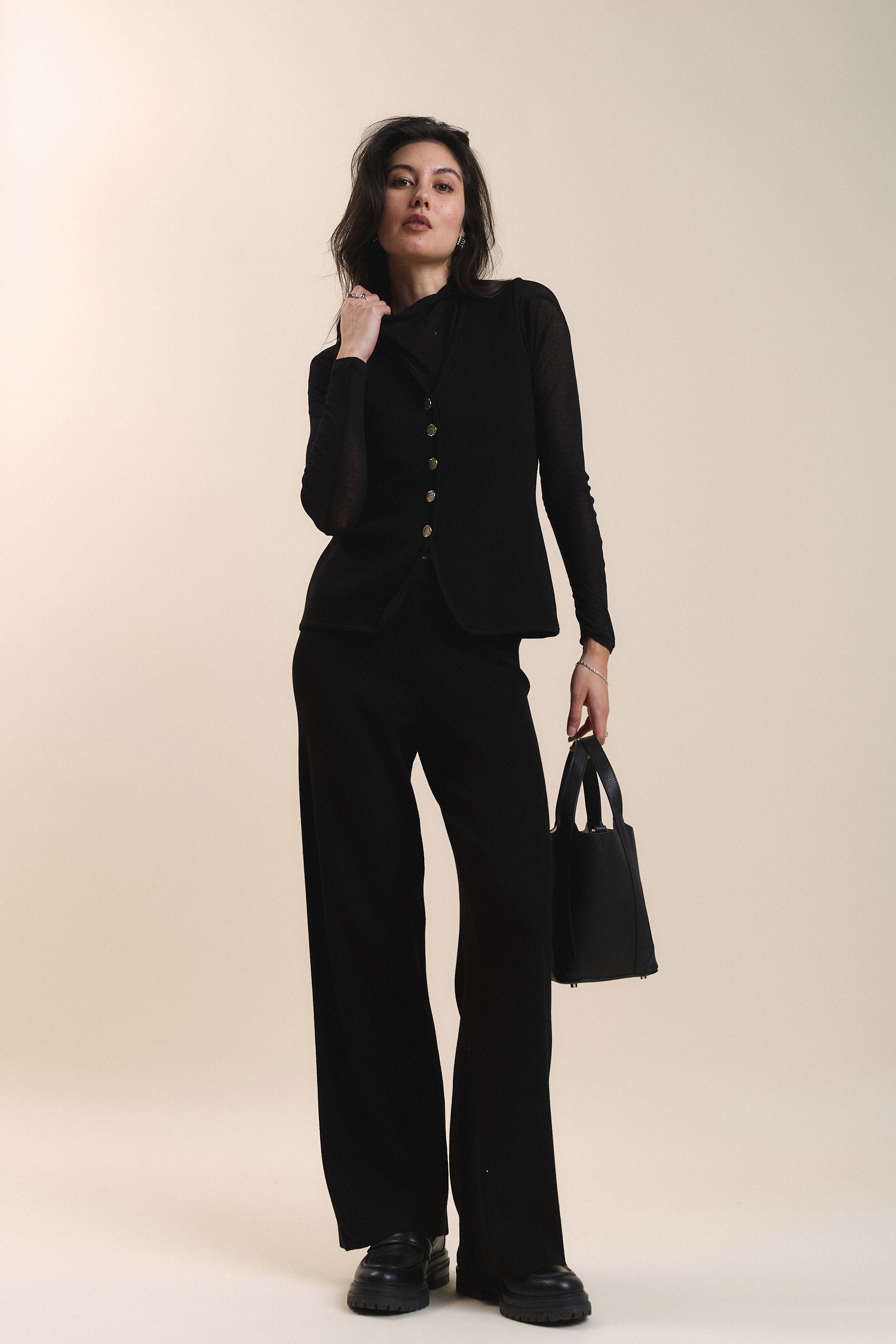 Woman in Max Charlie Knitted Pants holding a black handbag, standing by a beige background.