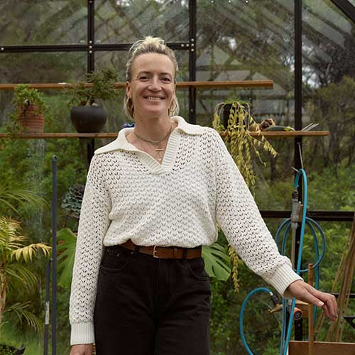 A smiling woman stands in a greenhouse, wearing a white knit sweater and black pants, surrounded by green plants and gardening tools.