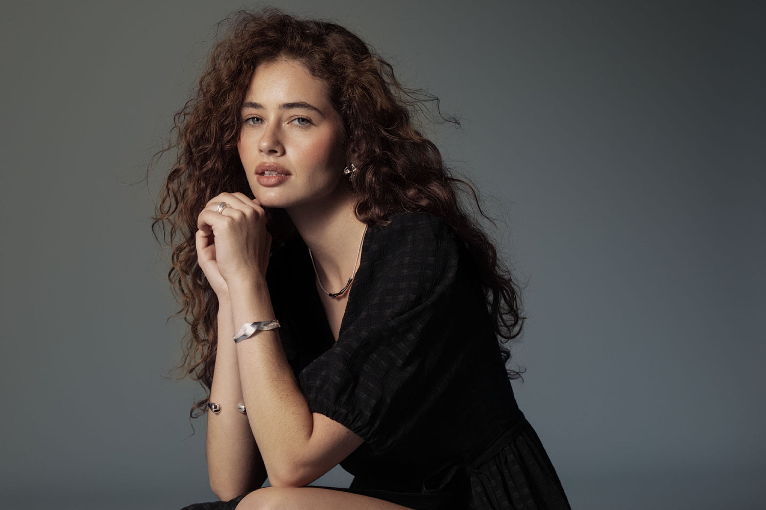 Woman with long curly hair in a black dress poses NZ style with hands clasped under chin.