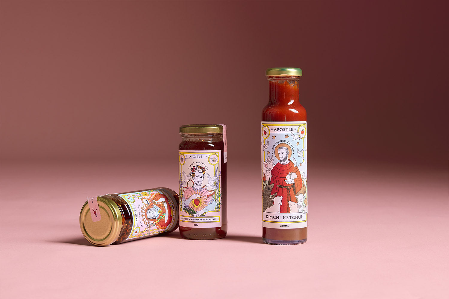 Three small jars of Apostle condiments with illustrated labels are stacked in front of a bowl of lemons and pears, placed on a kitchen countertop by a window.