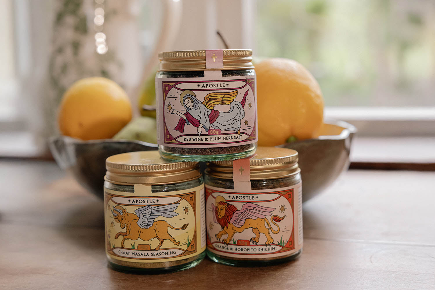 Three small jars of Apostle condiments with illustrated labels are stacked in front of a bowl of lemons and pears, placed on a kitchen countertop by a window.