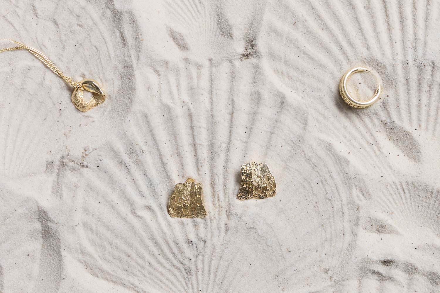 Gold jewelry, including a necklace, two earrings, and a ring, displayed on white sand with shell-shaped patterns in the background.