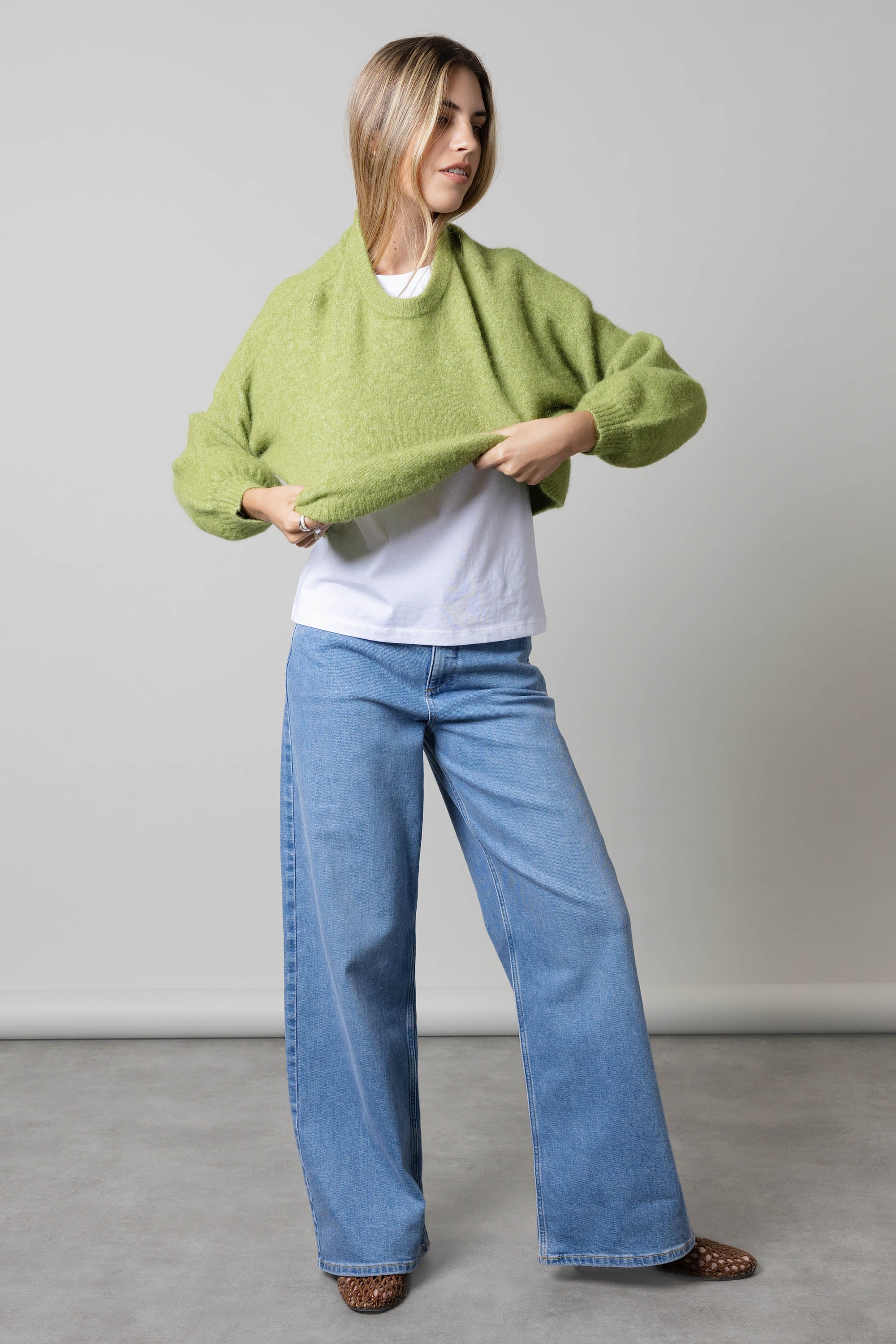 Woman in Nevada Wide Jeans by Max, green sweater, and white shirt on a gray floor.