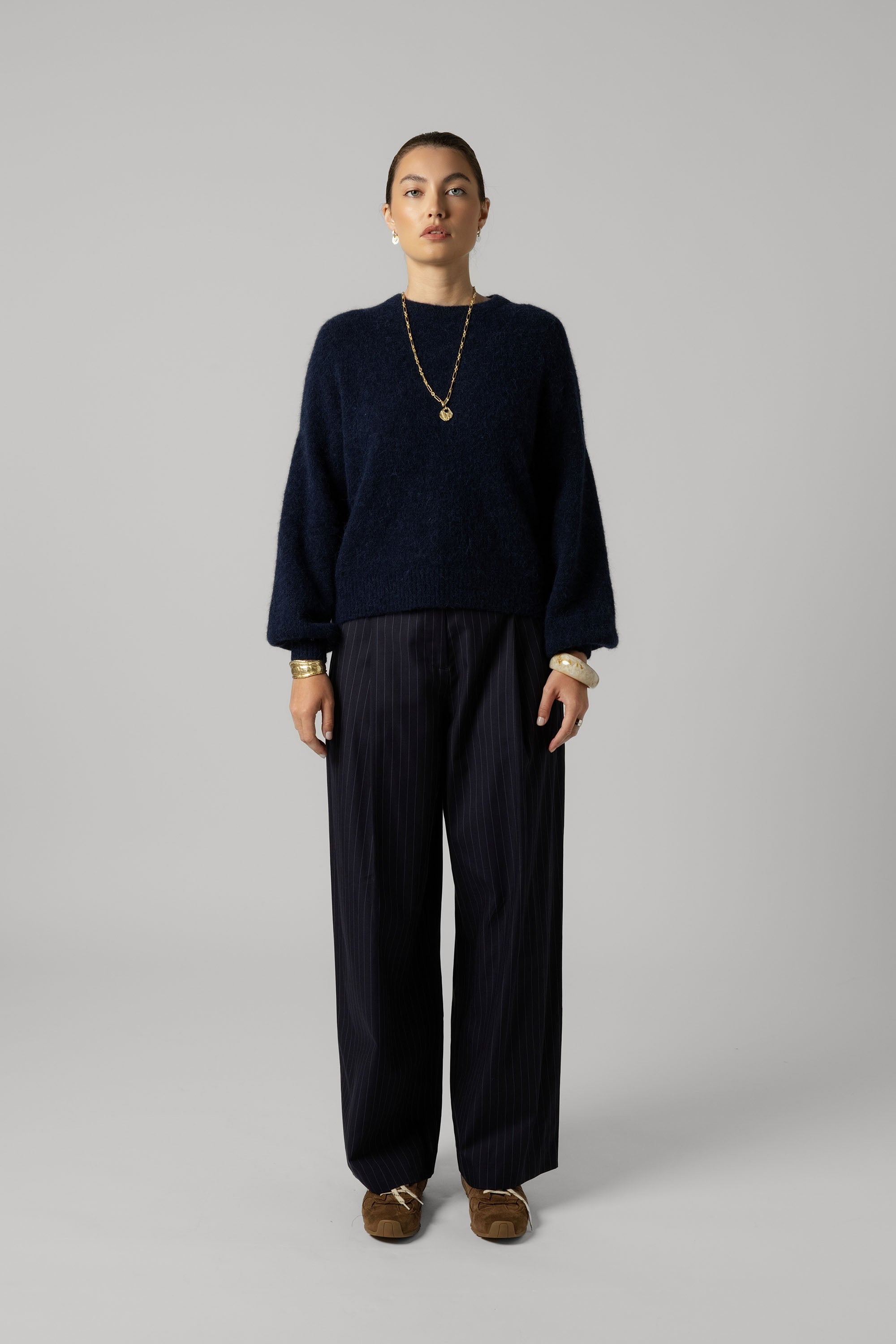 Woman wears Max's The Pleat Detail Wide Leg Pants in navy pinstripe with a dark sweater and gold jewelry.