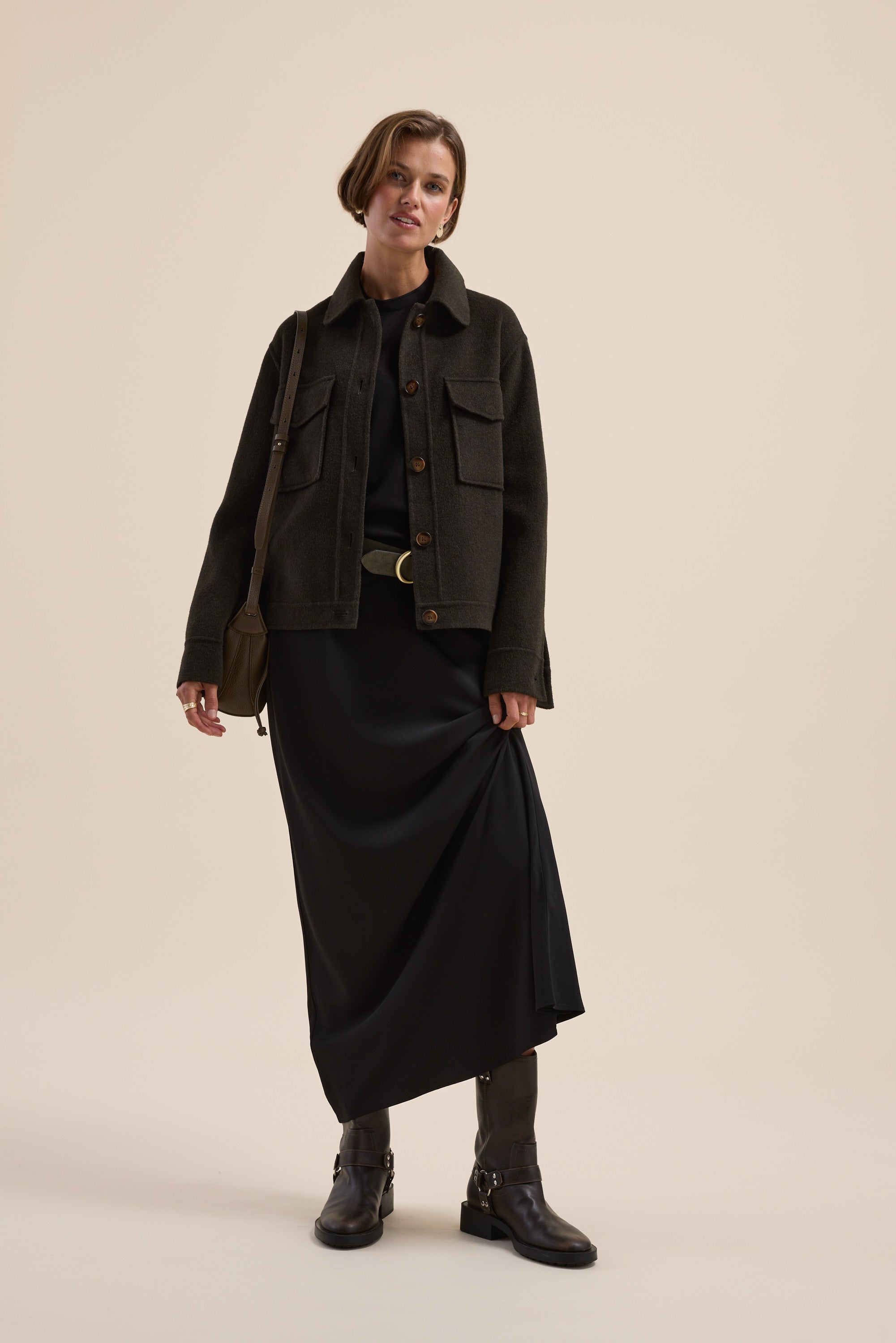 Woman in The Maxi Slip Skirt by Max poses against a beige background in boots and a dark jacket.