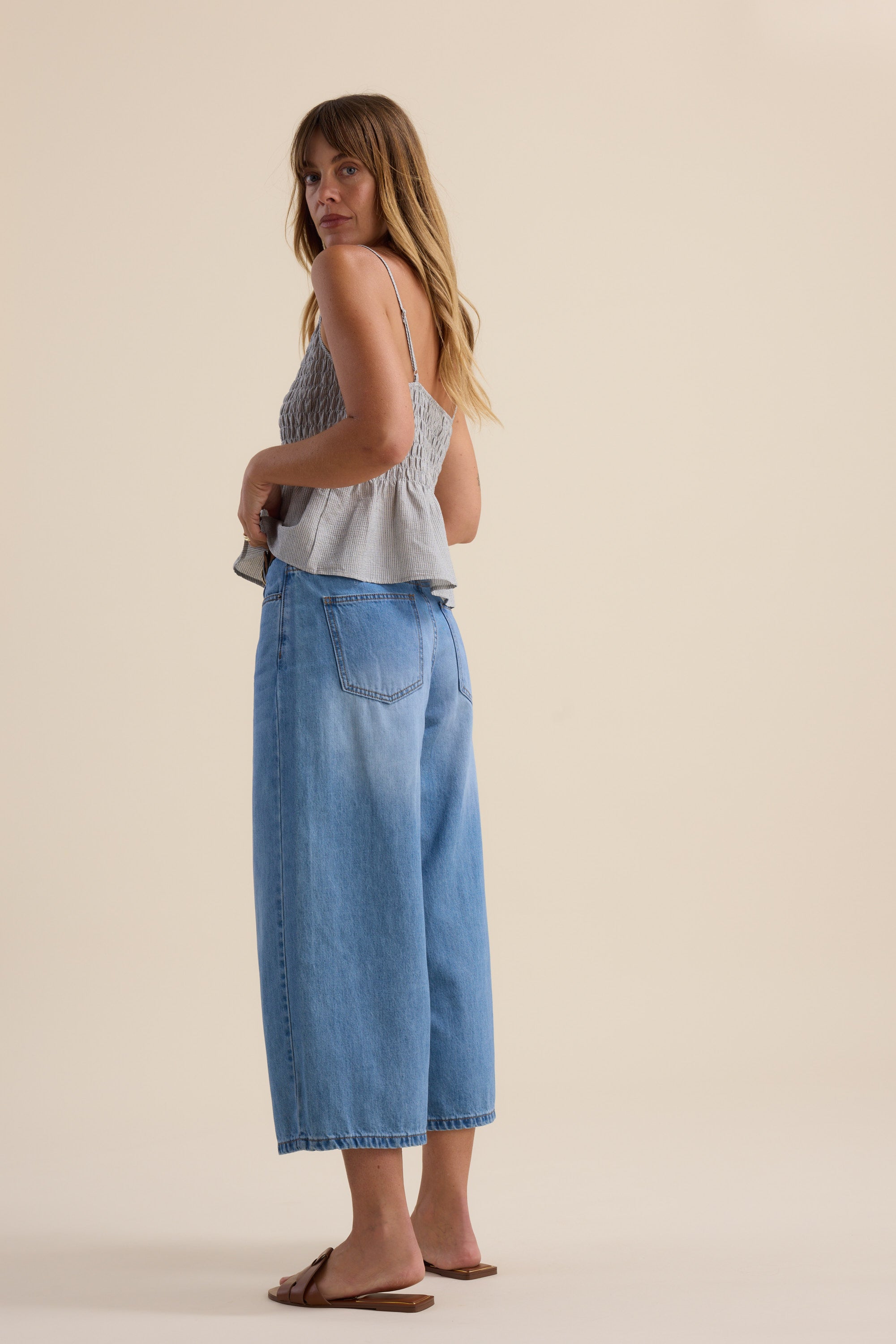 A woman stands barefoot on a light floor, wearing a gray smocked spaghetti strap top and Max's Derby Wide Leg Cropped Jeans. She is turned slightly sideways, looking over her shoulder.