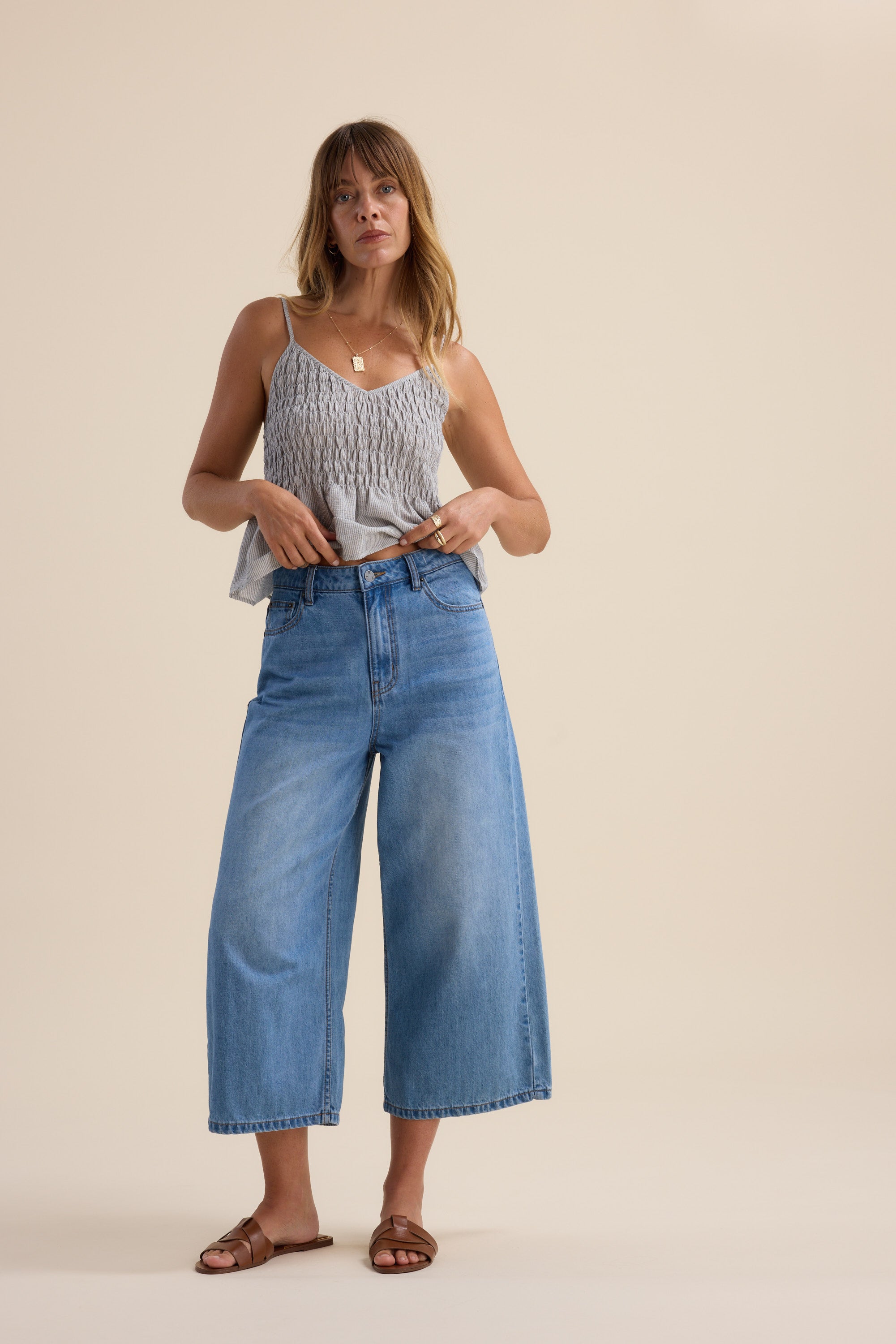 A woman stands against a plain background wearing Max's Derby Wide Leg Cropped Jeans, a light gray sleeveless top, brown sandals, and a gold necklace. She holds her high rise jeans at the waist and looks forward with a neutral expression.