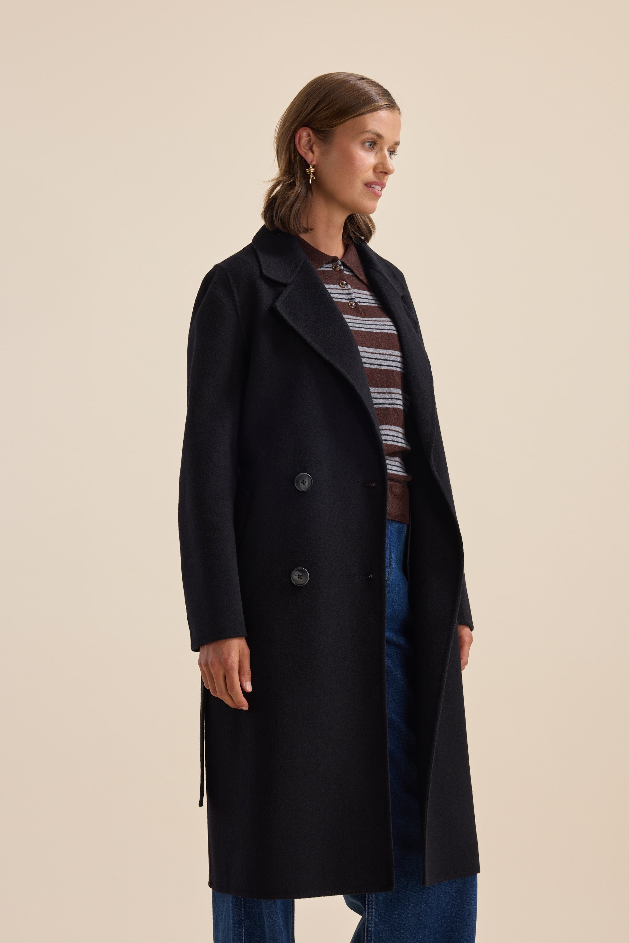 Woman in The Belted Wool Coat by Max, striped top, and blue jeans stands against beige background.