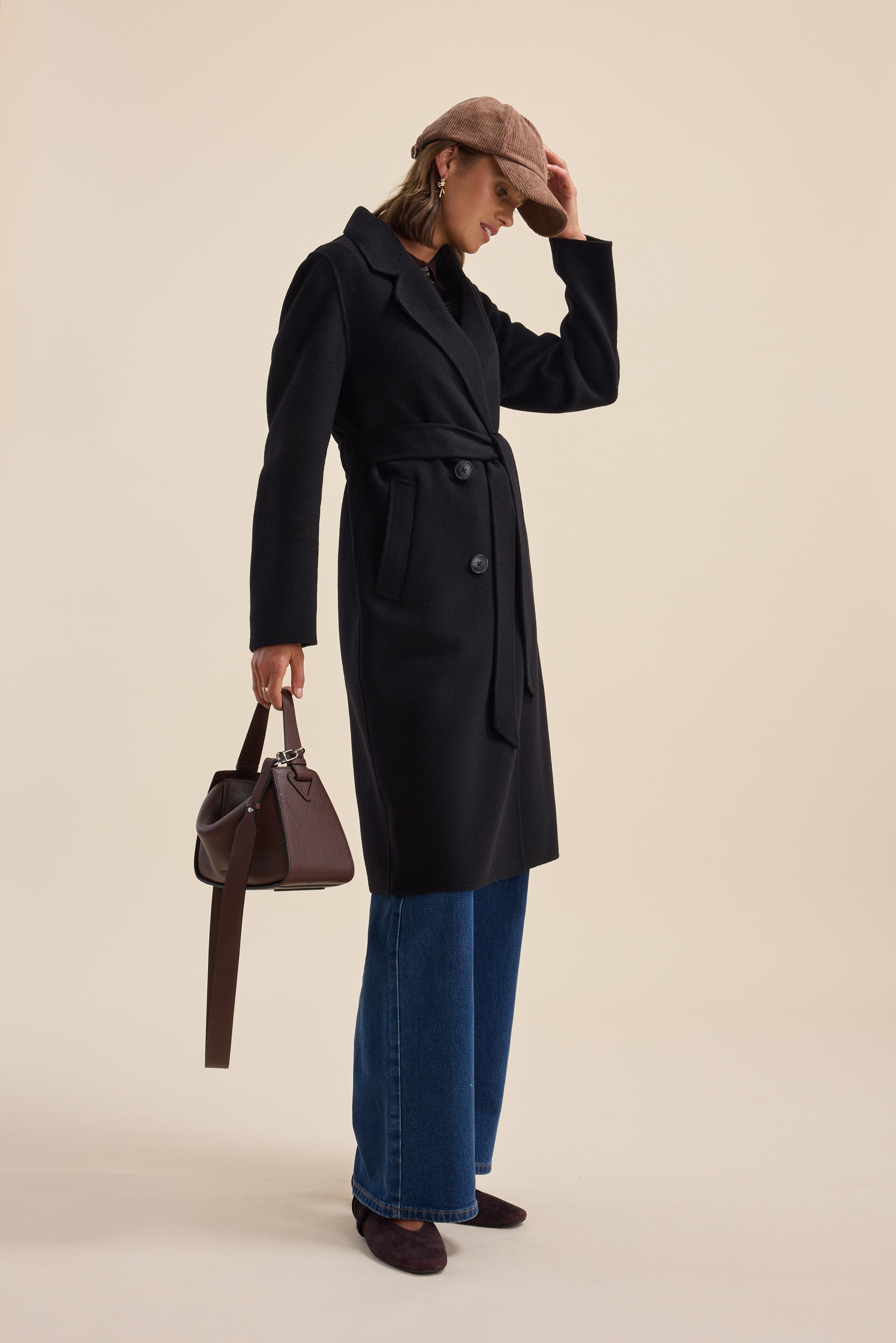 Woman wearing The Belted Wool Coat by Max, blue jeans, brown hat, and holding a brown handbag.