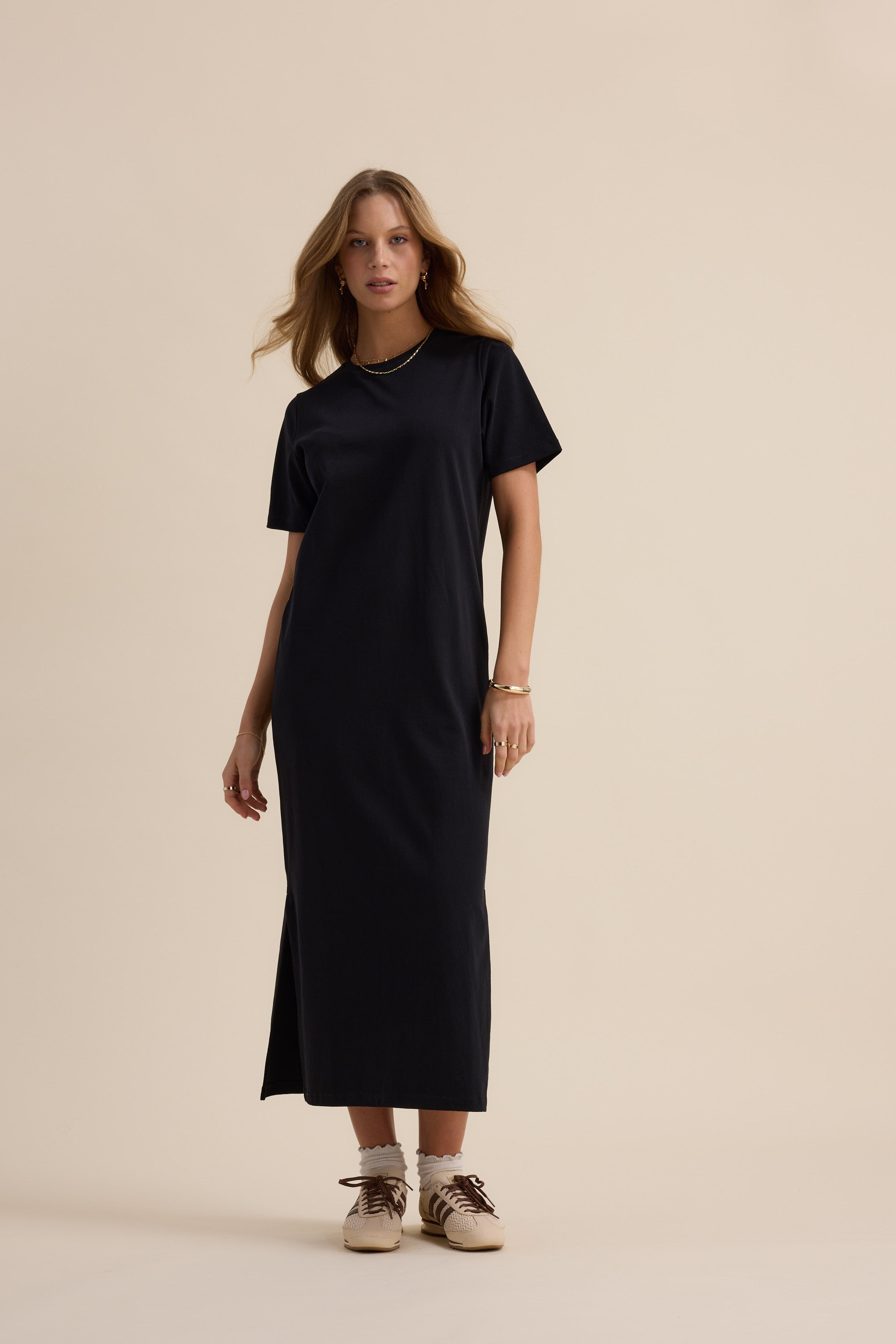 Woman wears The Tee Shirt Dress by Max with sneakers, standing against a beige background.