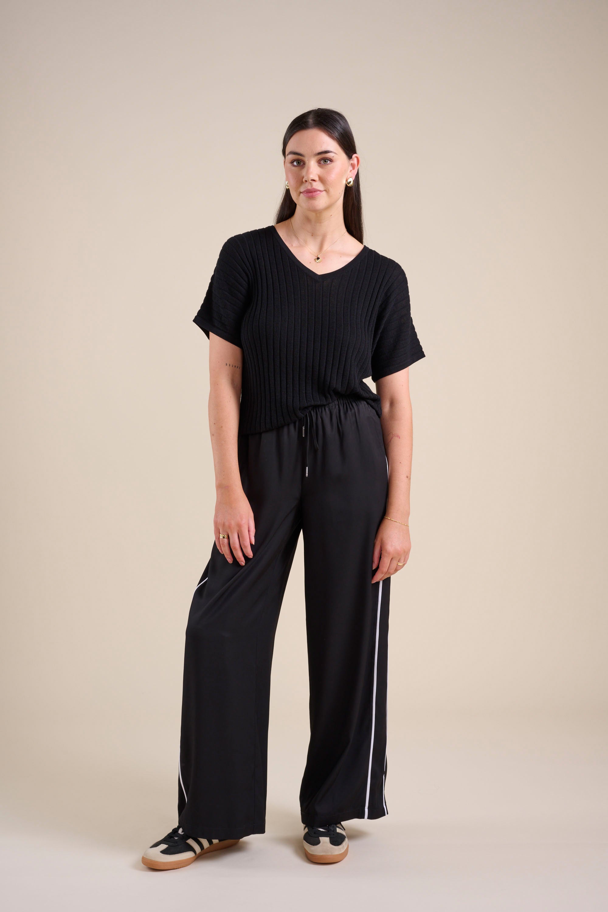 A woman with straight dark hair and a neutral expression wears Max's The Elevated Jogger—wide-leg striped joggers with white side stripes—paired with a black short-sleeved knit top and black sneakers, standing against a plain beige background.