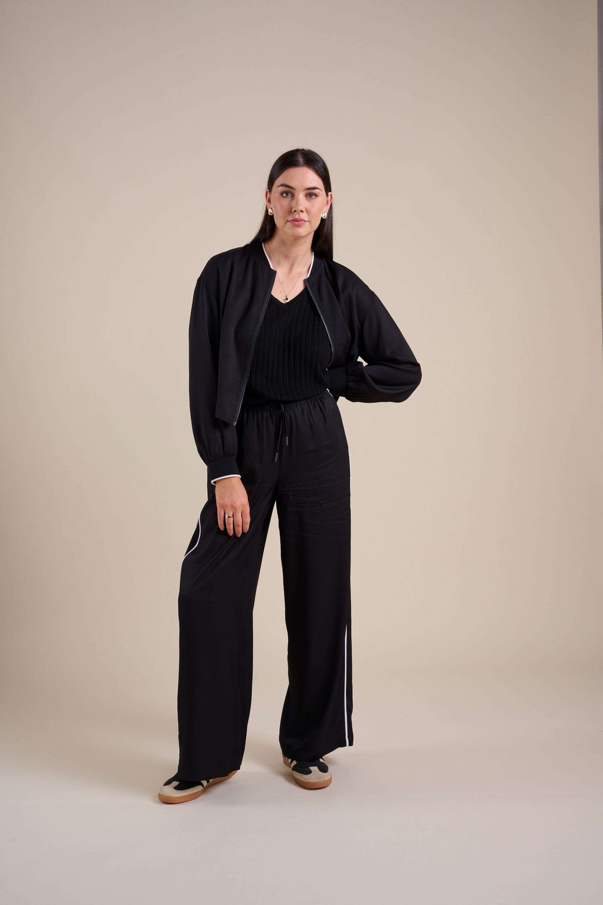 A woman with straight hair and a neutral expression stands against a beige background, wearing Max's The Elevated Jogger—high-rise wide leg jogger pants with white side stripes—paired with a black jacket and top, one hand on her hip.