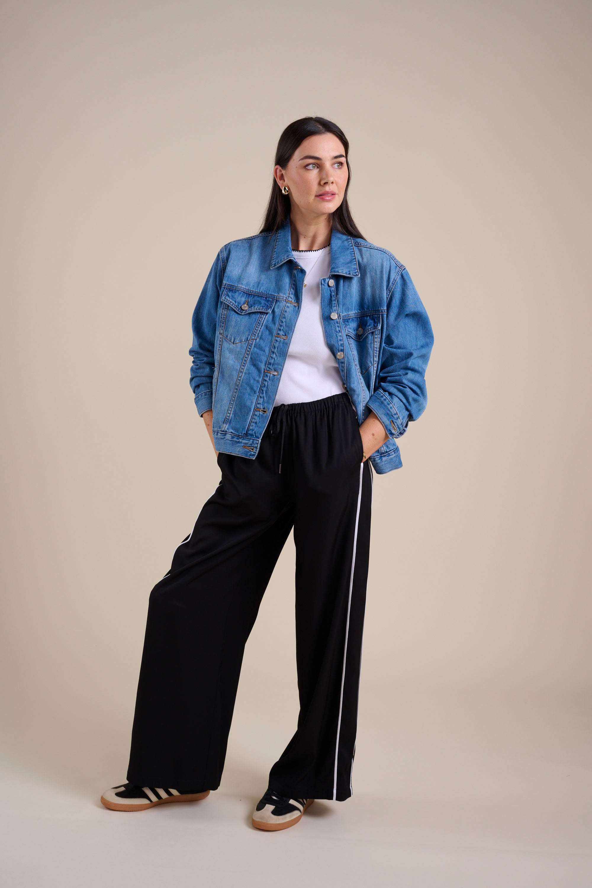 A woman stands with hands in pockets, wearing Max's The Elevated Jogger—black high rise pants with a white stripe—paired with a blue denim jacket, white t-shirt, and black sneakers, set against a plain beige background.