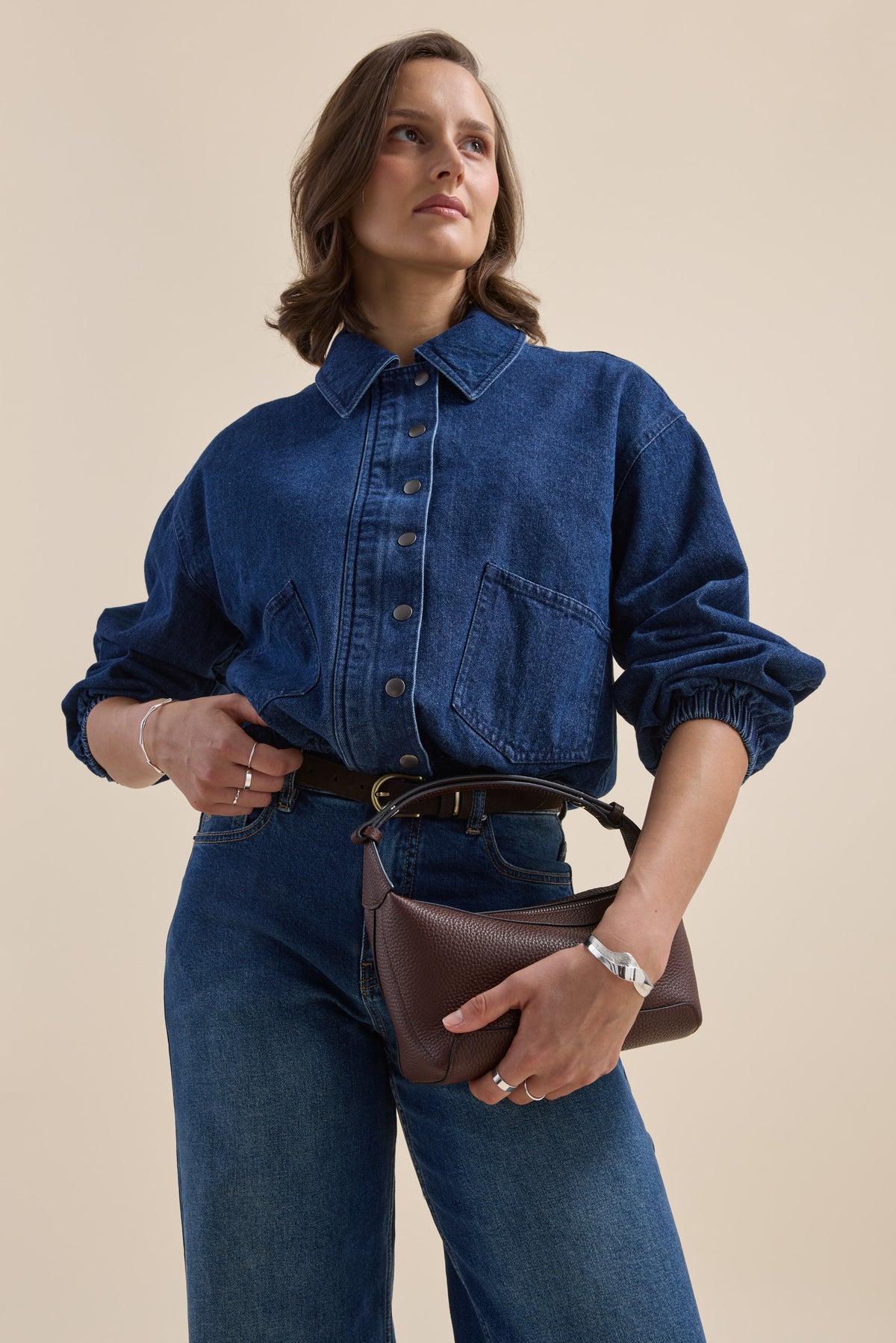 Woman in Elementary Relaxed Jacket and jeans, holding a brown purse, beige background.