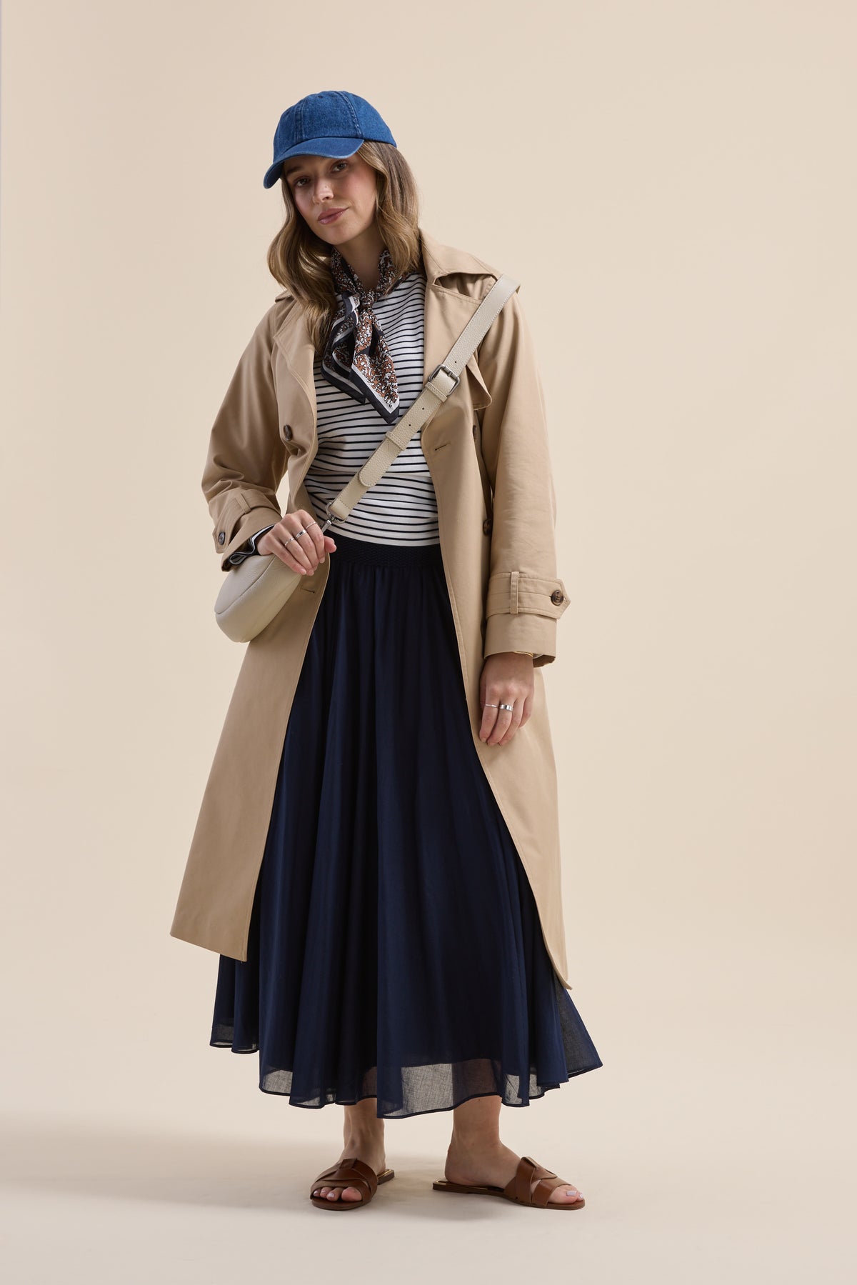 Woman in trench coat, striped top, Elementary Gathered Skirt, blue cap, sandals, crossbody bag.