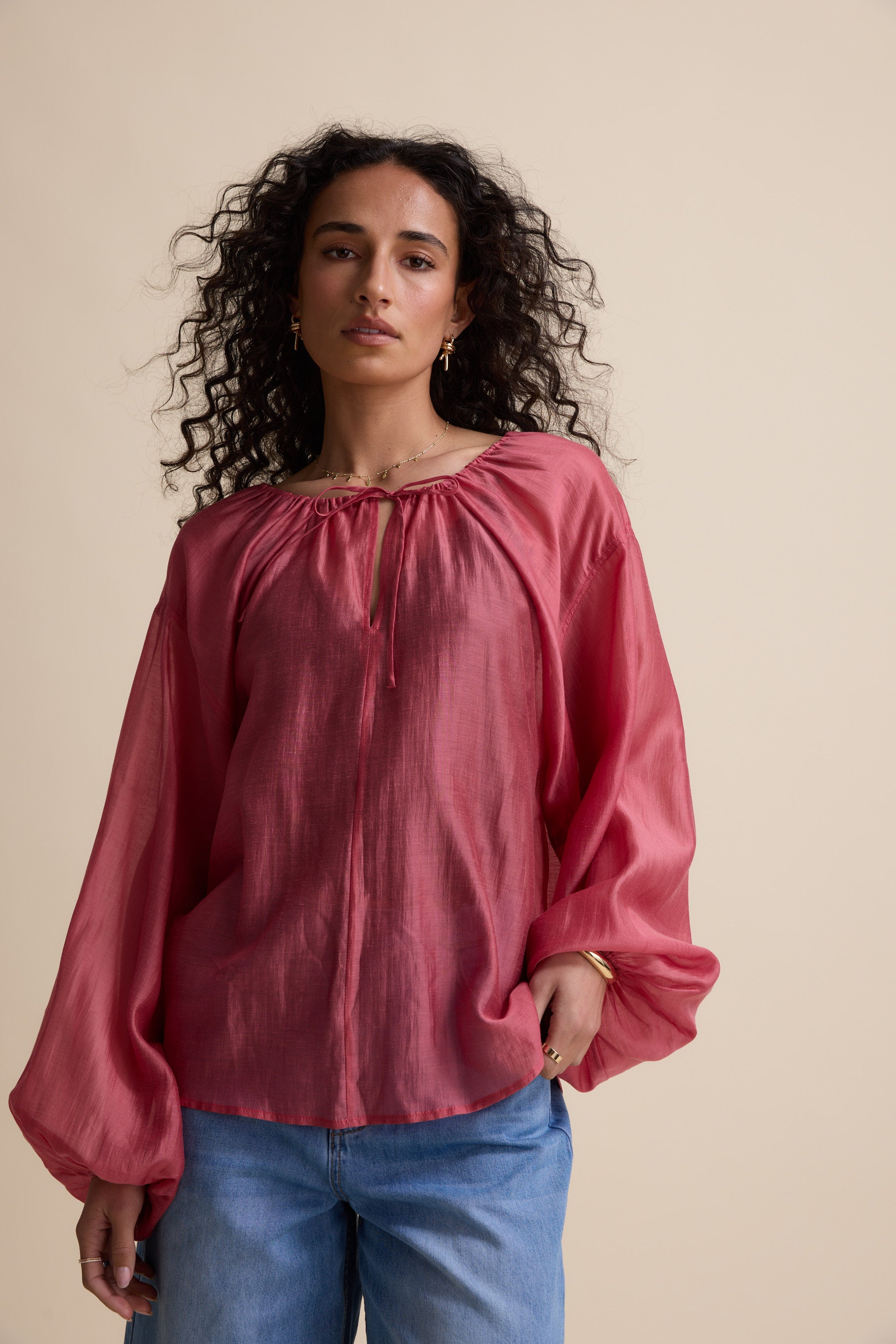 Woman with curly hair in Max’s Abigail Tie Neck Blouse and blue jeans stands by a beige background.