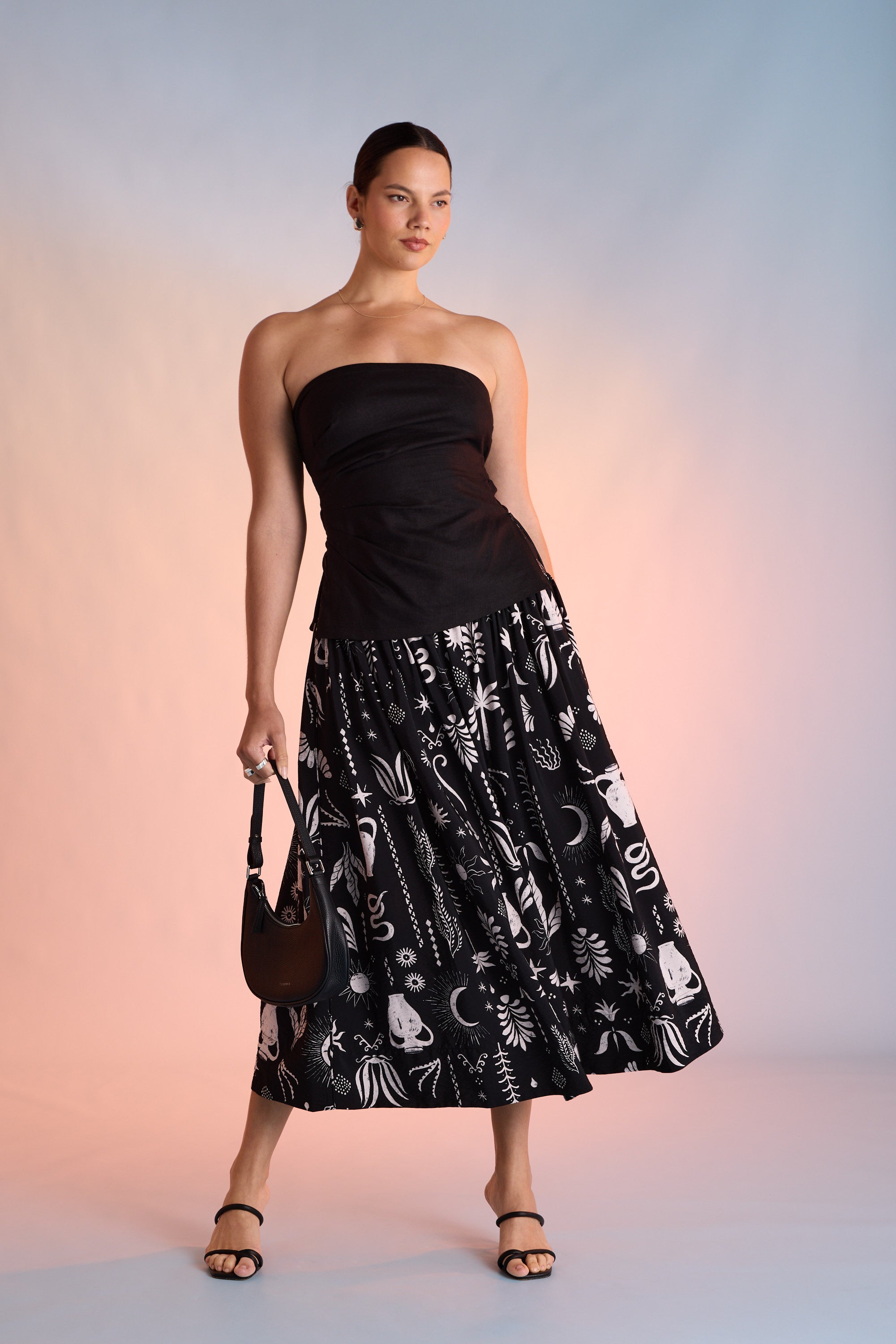 A woman exudes confidence in the Max Helena Drop Waist Skirt with an exclusive print, paired with a strapless black top, black sandals, and a chic handbag, posing against a soft gradient background.