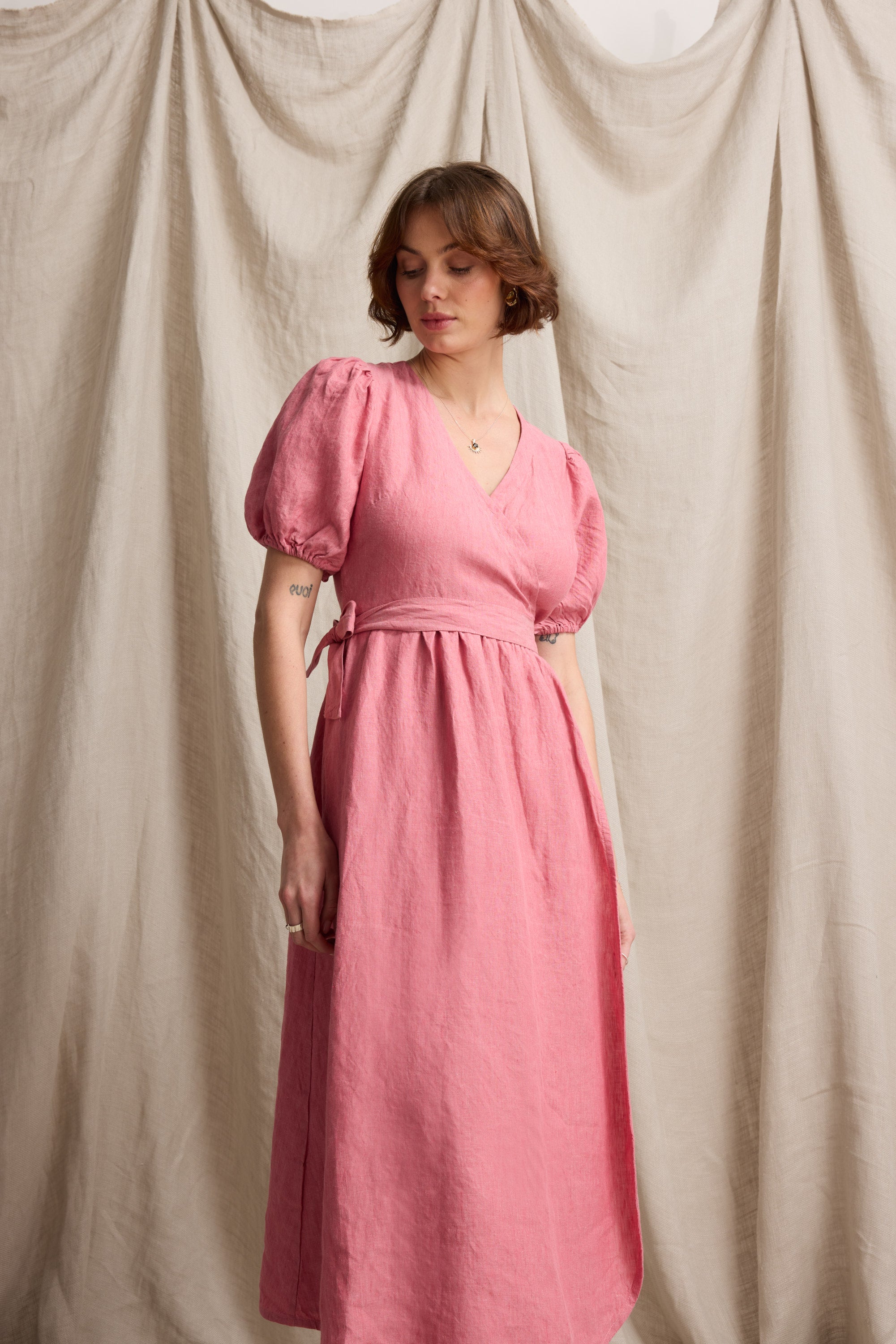 Woman in the Max Sara Linen Wrap Dress stands before beige draped fabric, looking down.