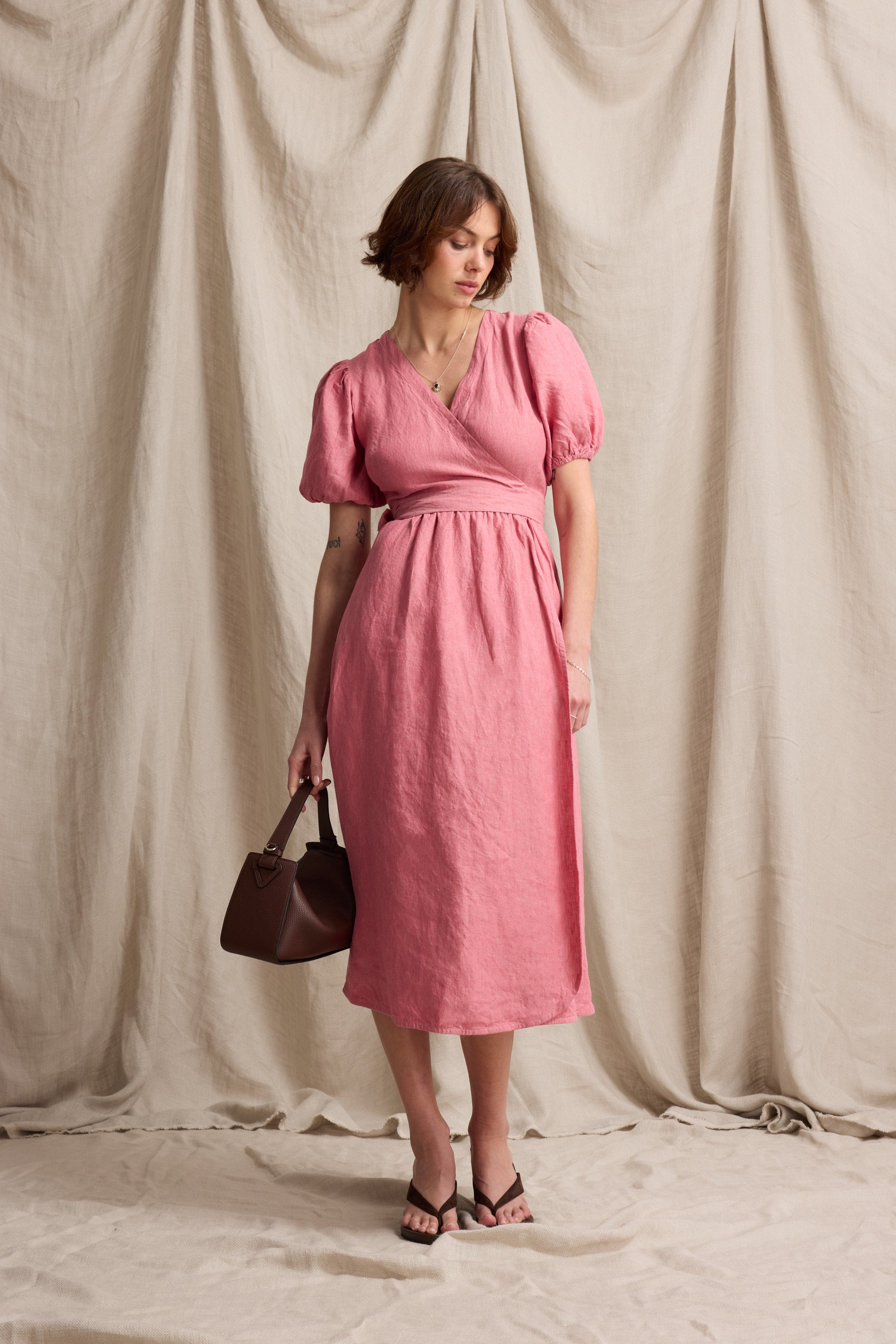 Woman in the Sara Linen Wrap Dress by Max holds a brown bag, posed against a beige backdrop.