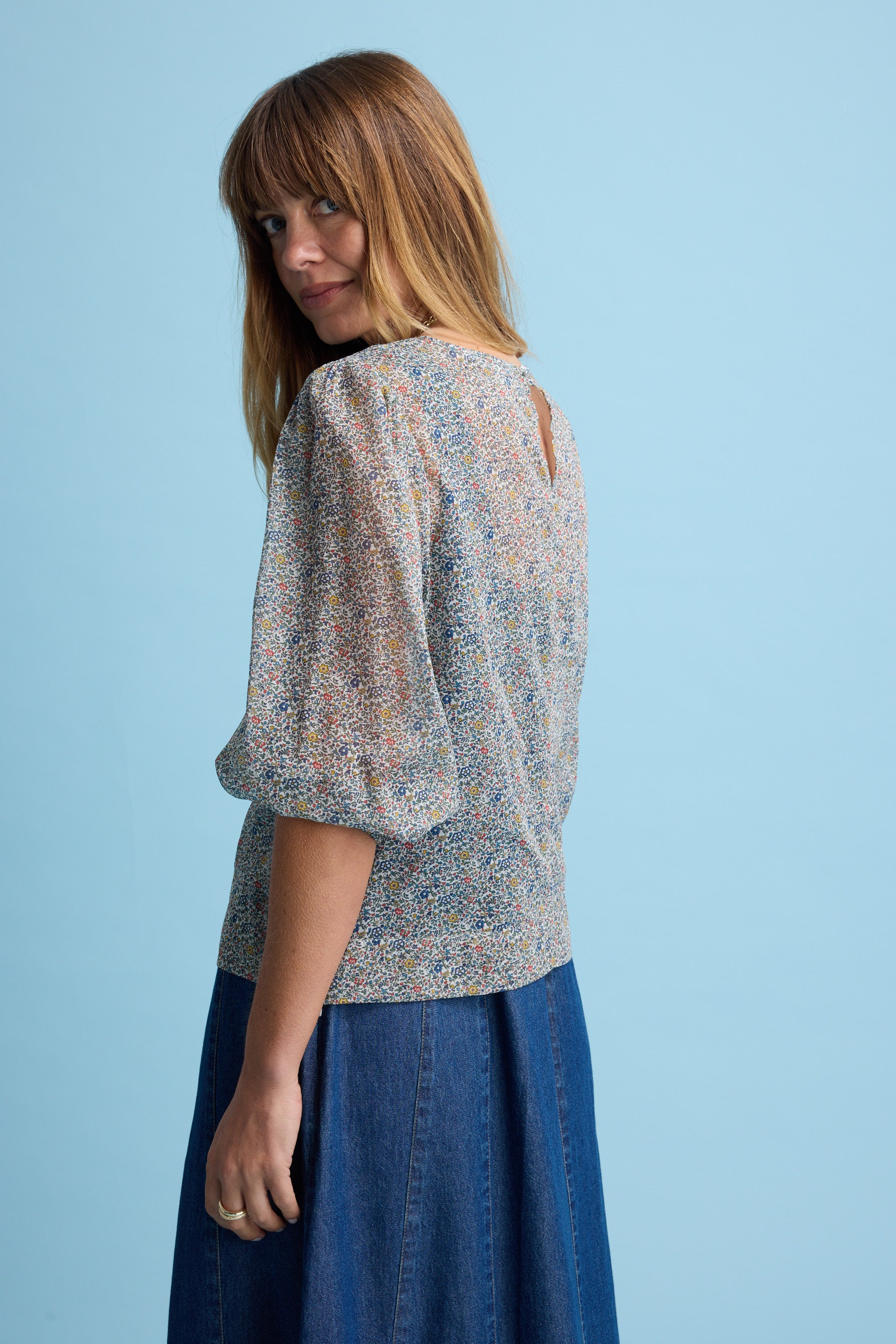 A woman with straight light brown hair and bangs poses turned slightly away, looking over her shoulder. She wears the Eloise Puff Sleeve Blouse by Max and a blue denim skirt against a light blue background.