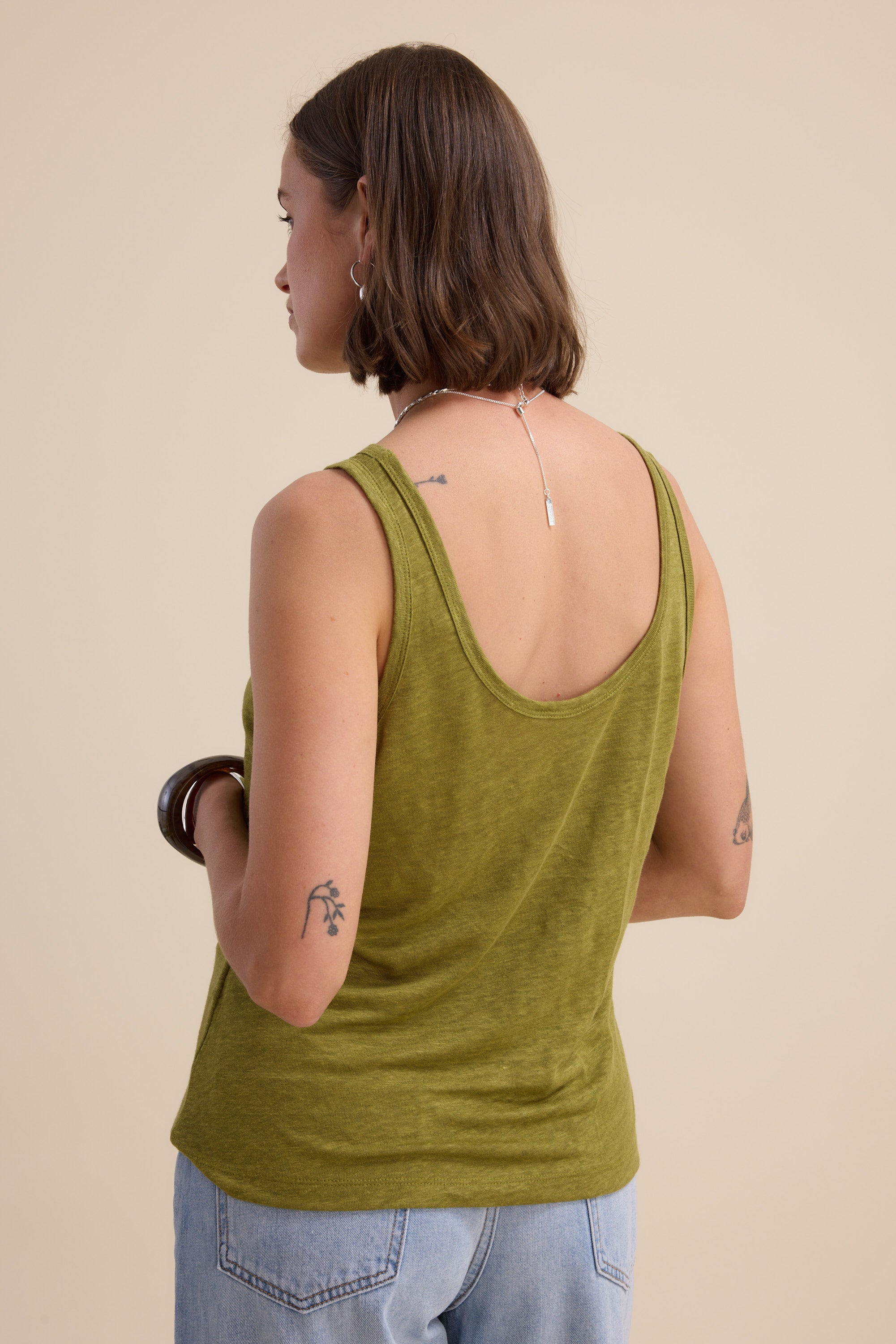 A person with short brown hair faces away, revealing small arm tattoos, while wearing the Elementary Linen Jersey Tank from Elementary in green and light blue jeans, set against a plain beige background.