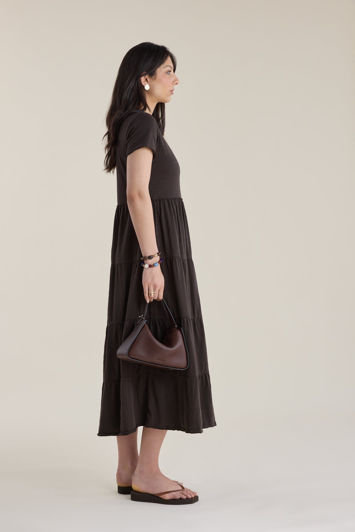 Woman in Elementary Jersey Tiered Dress, holding a handbag, stands sideways before plain backdrop.