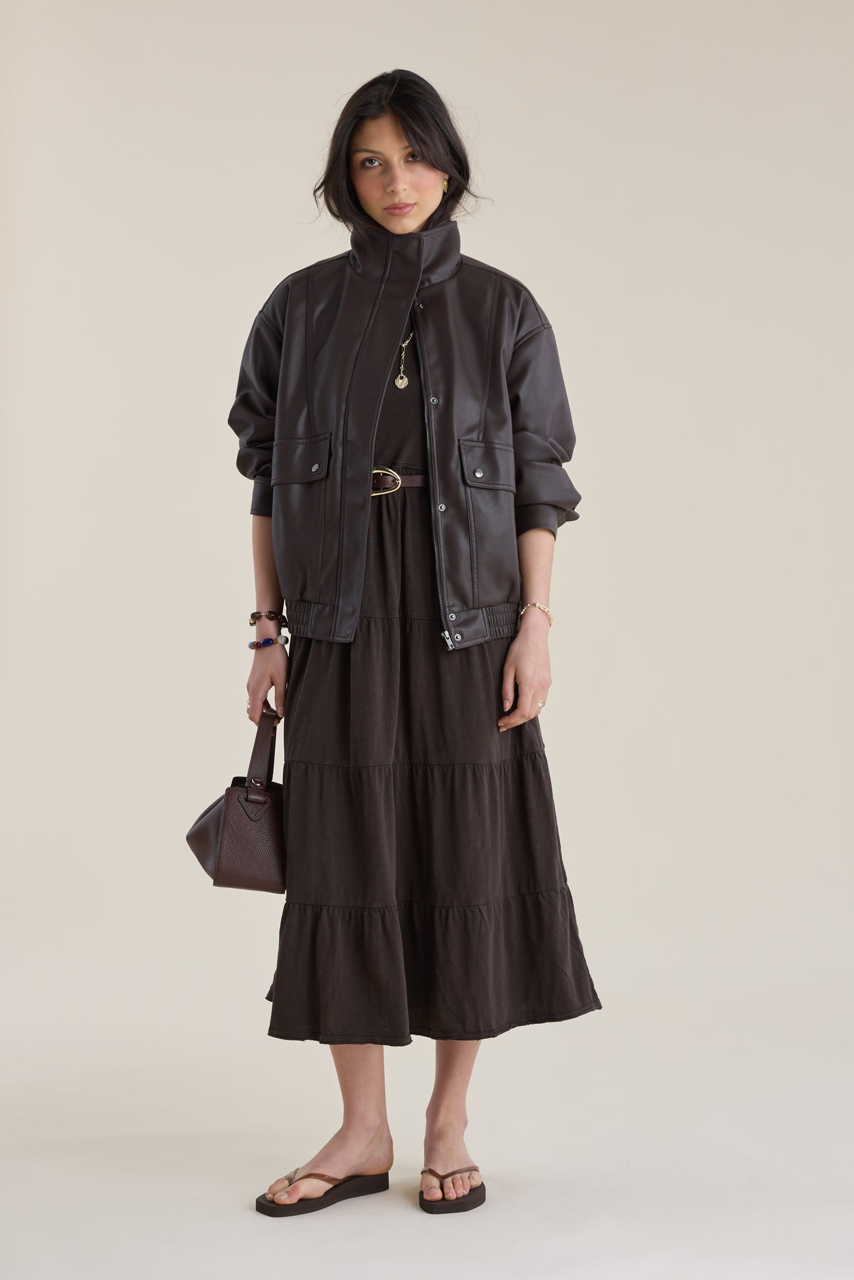 A woman wears an Elementary Jersey Tiered Dress, dark jacket, sandals, and holds a matching bag.