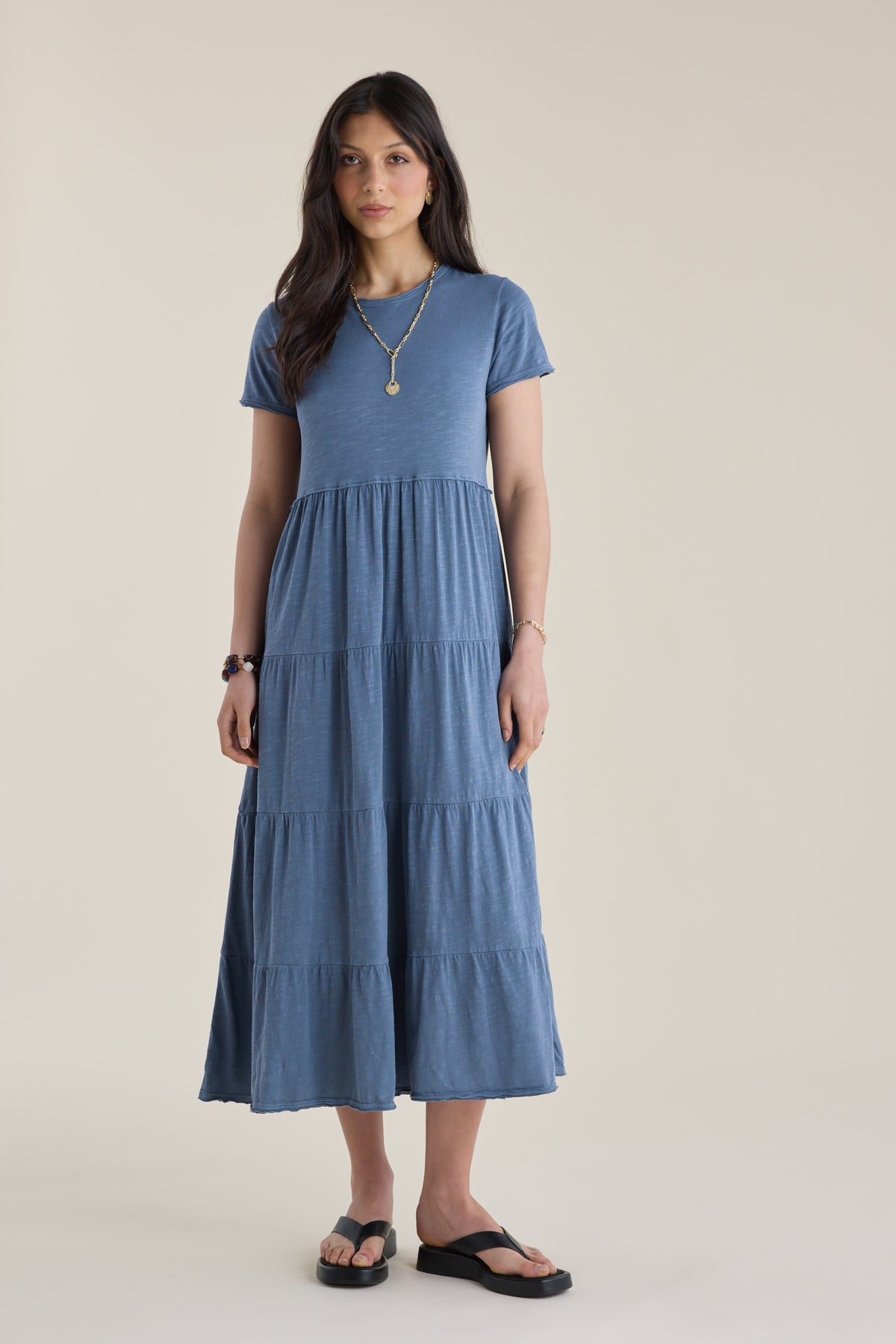 Woman in Elementary Jersey Tiered Dress, black sandals, and necklace stands on a light floor.