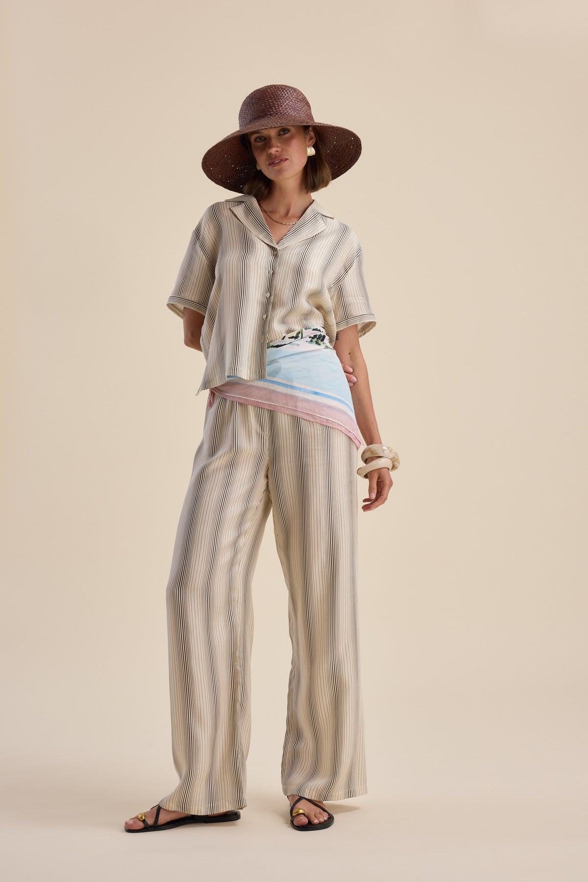 Woman in Max Sera Drawstring Pants, sunhat, sandals, and pastel scarf against beige backdrop.