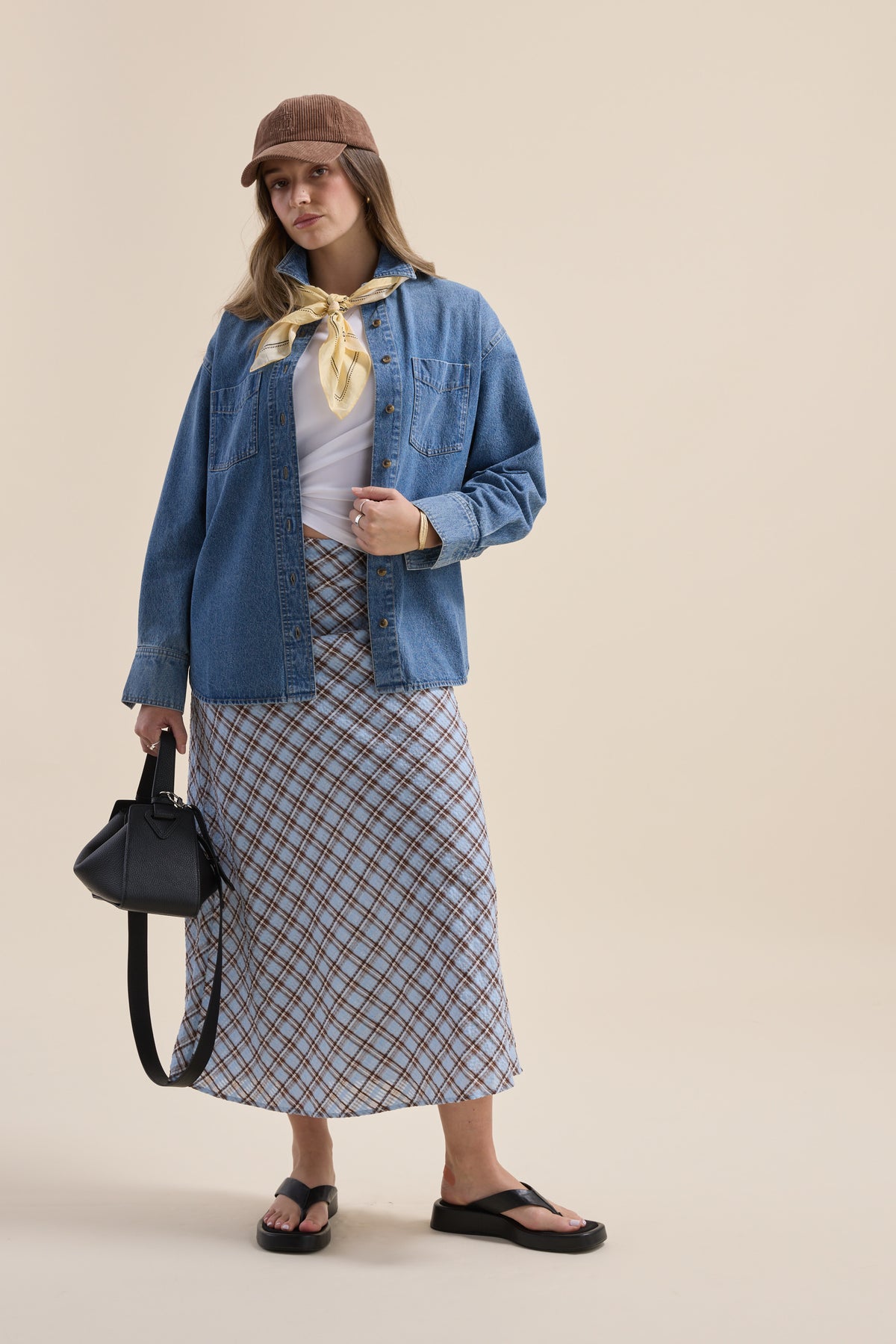 Woman in denim shirt, The Perfect Slip Skirt by Max, yellow scarf, brown cap, black bag, sandals.