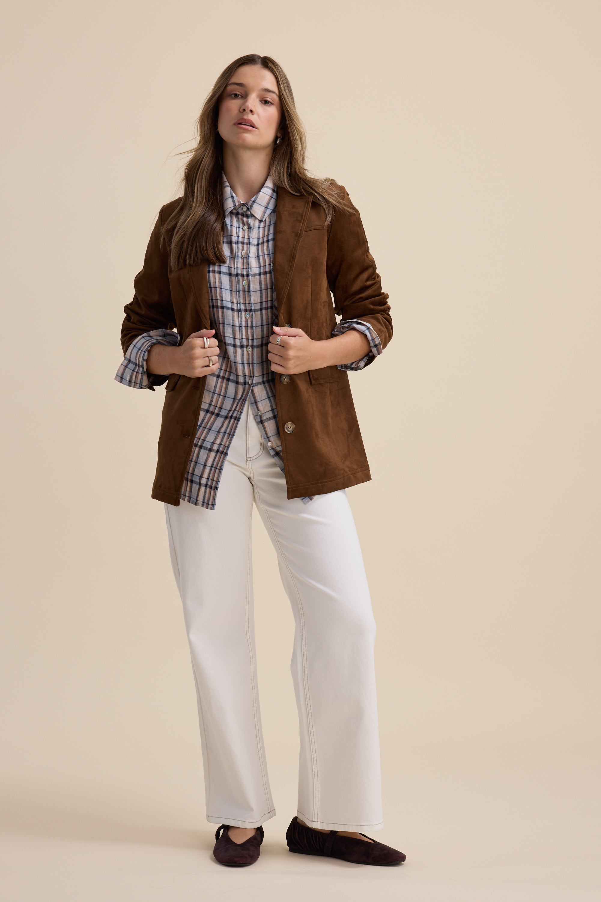 Woman in brown jacket, Elementary Oversized Shirt, white pants, and dark shoes on beige background.