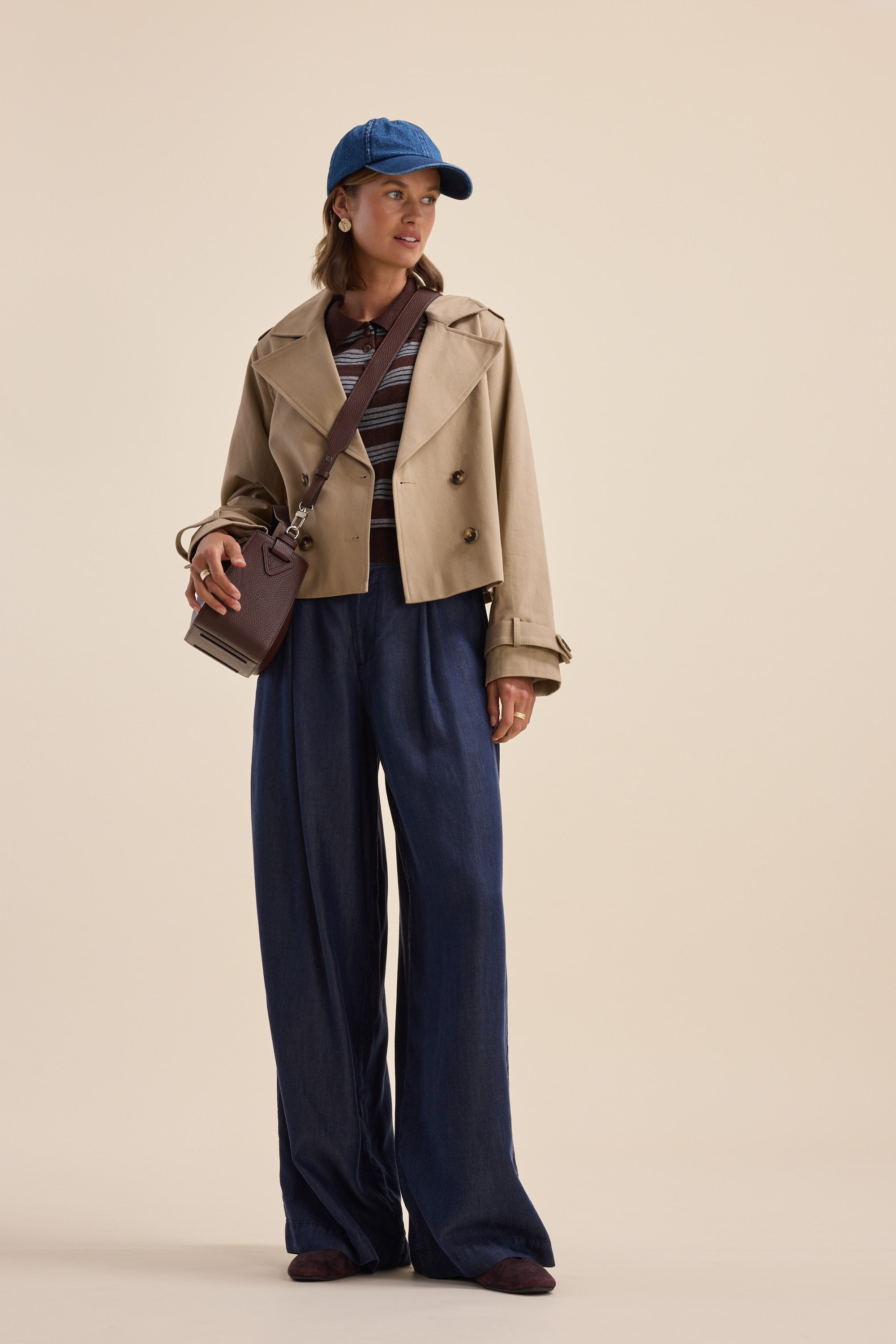 Woman in tan jacket, Elementary Henley tee, wide-leg jeans, blue cap, and crossbody bag poses.