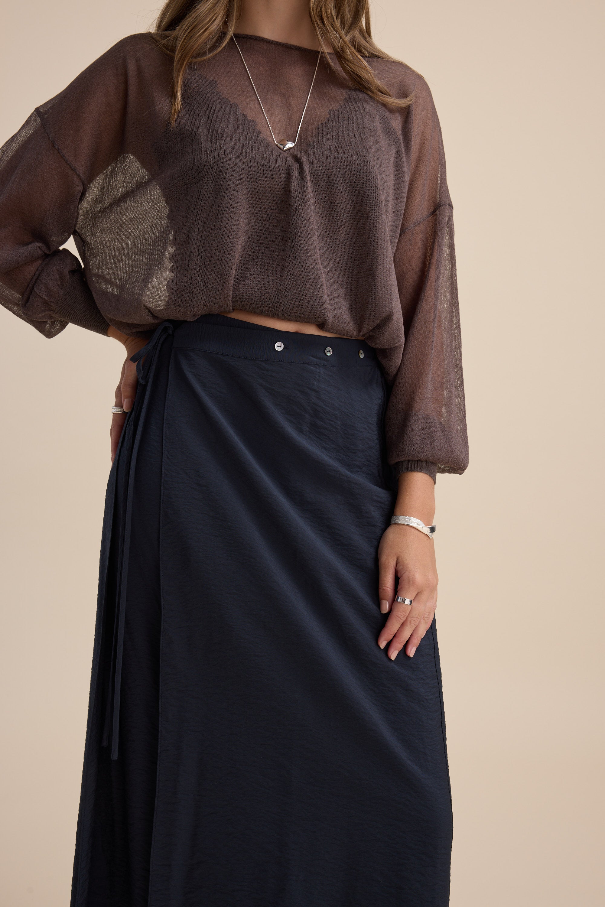 Woman in a sheer brown top and Elementary navy wrap skirt with jewelry and natural nails.