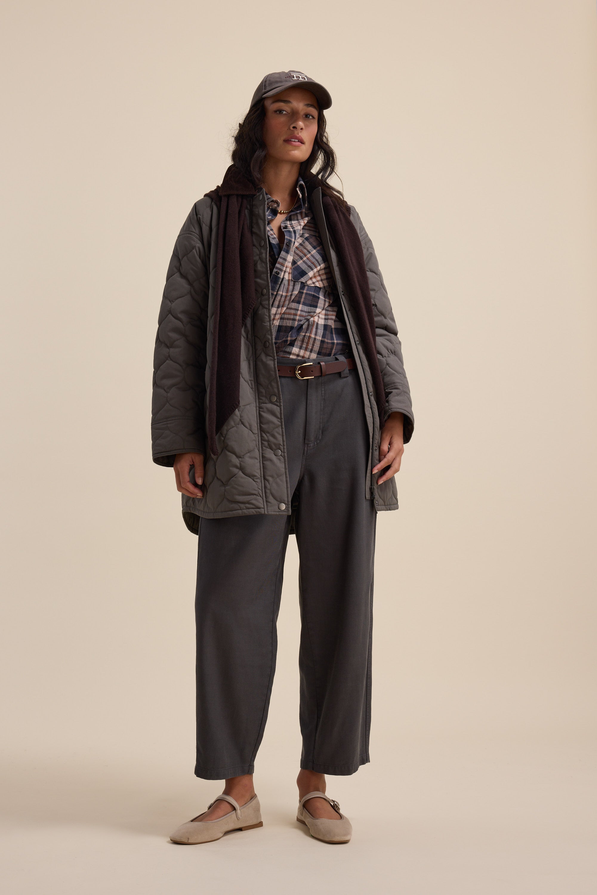 Woman in a grey quilted coat, plaid shirt, Elementary Panelled Barrel Pants, loafers, and a cap.