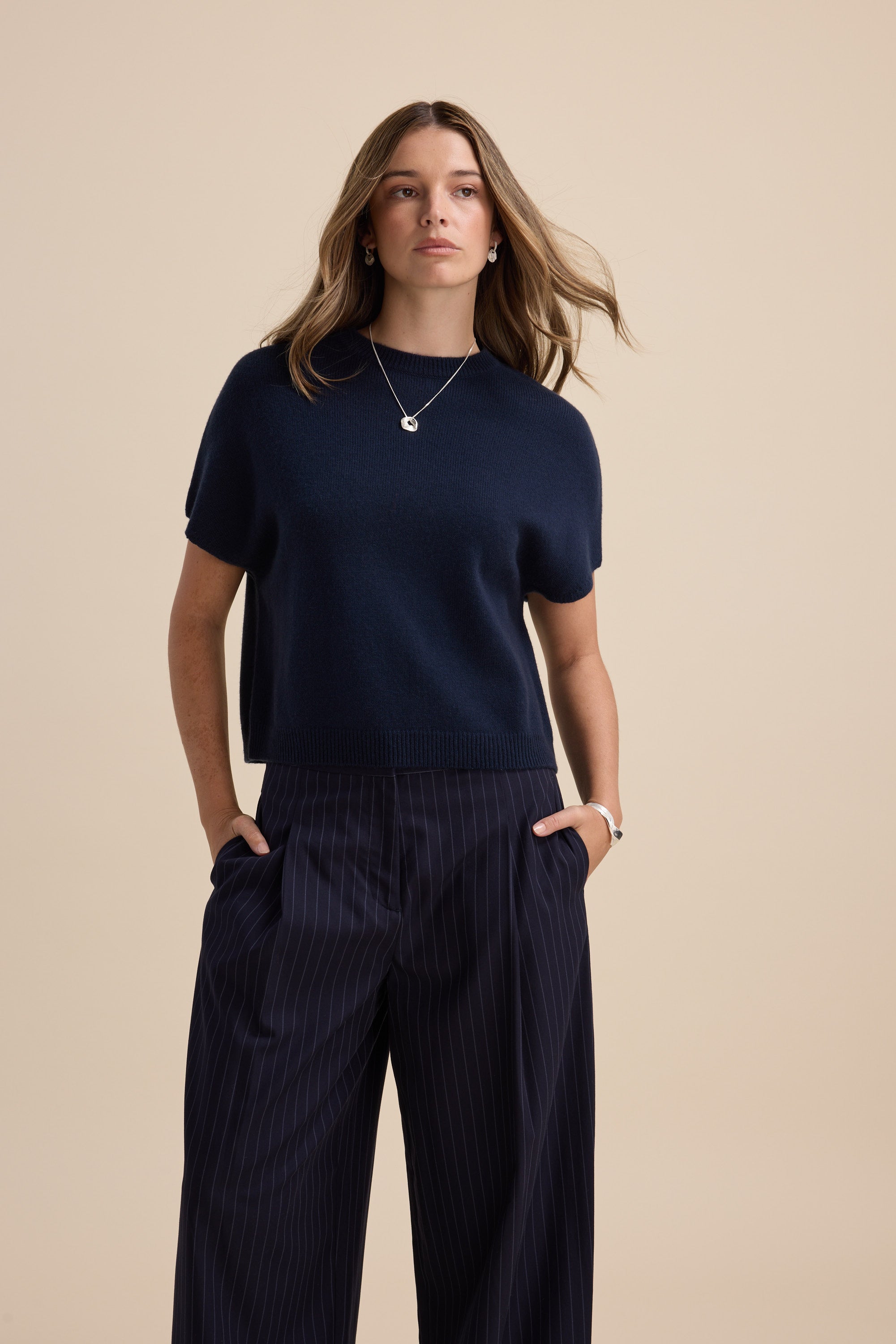 Woman in Elementary cashmere blend drop shoulder top and pinstripe pants against beige backdrop.