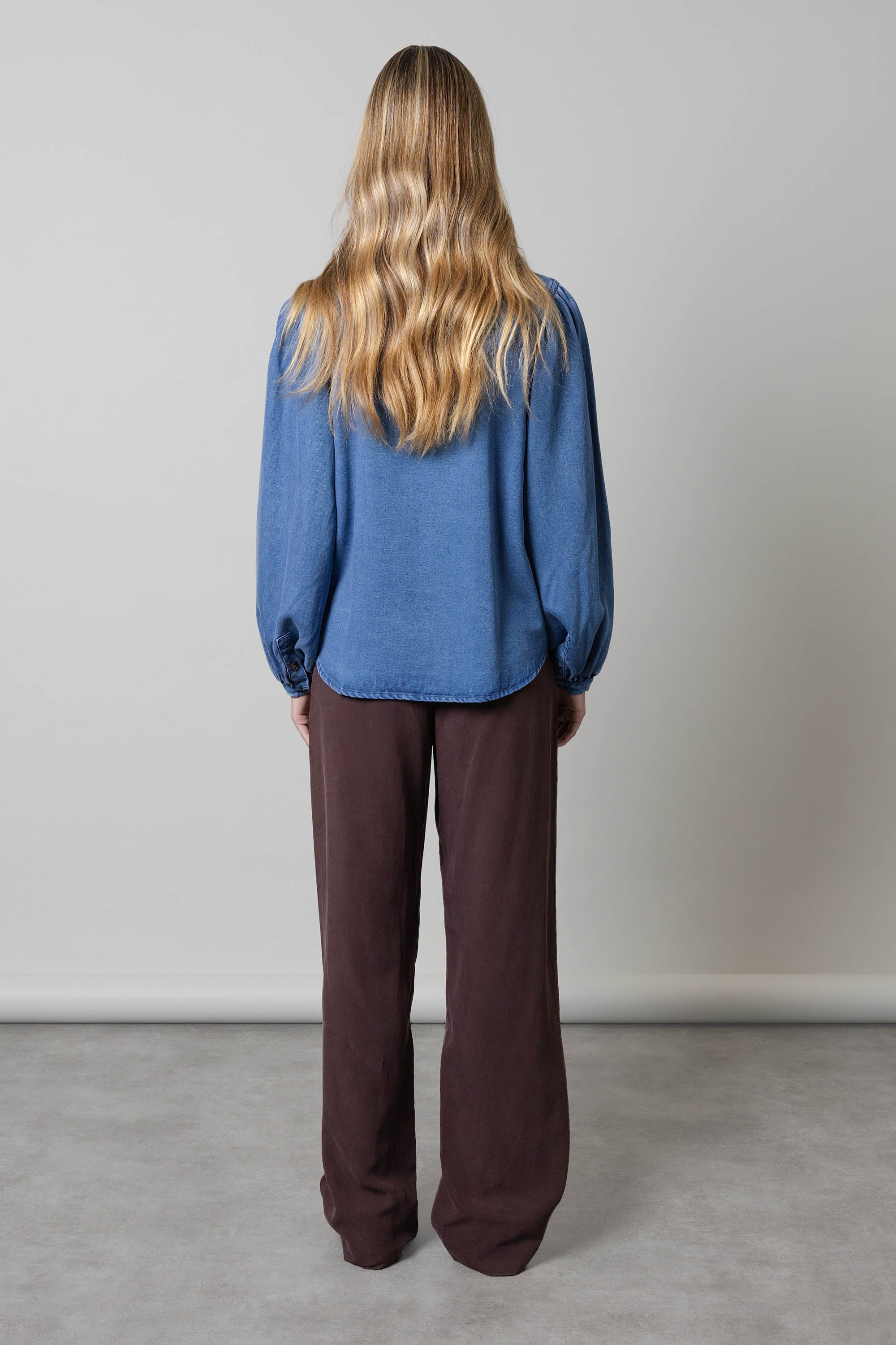 Person with long wavy hair in an Elementary Denim Shirt, standing facing away.