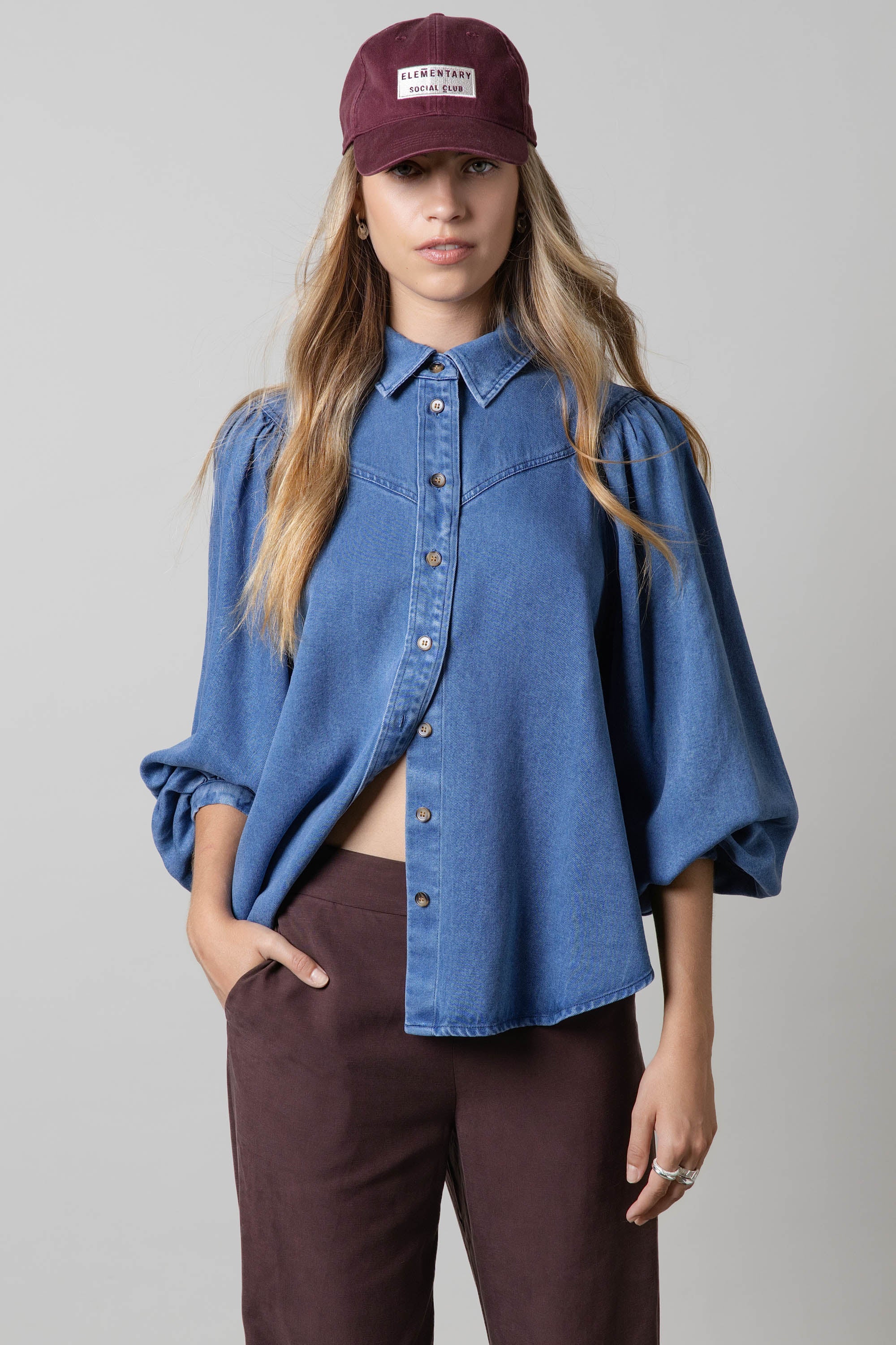 Woman in the Elementary Denim Shirt by Elementary, featuring blouson sleeves, and a maroon cap.