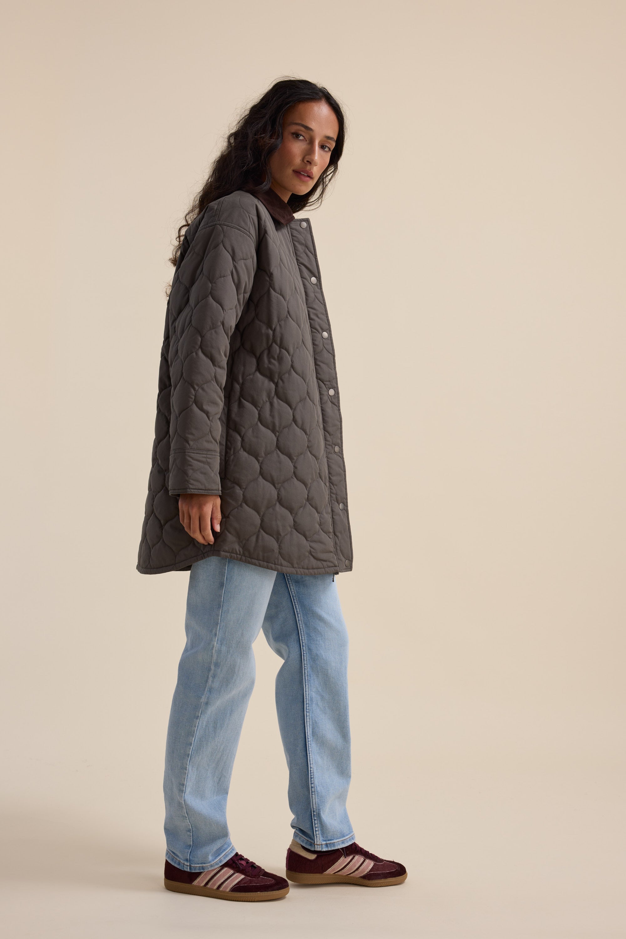 Woman in the Elementary Longline Puffer, light blue jeans, and maroon sneakers, side view.