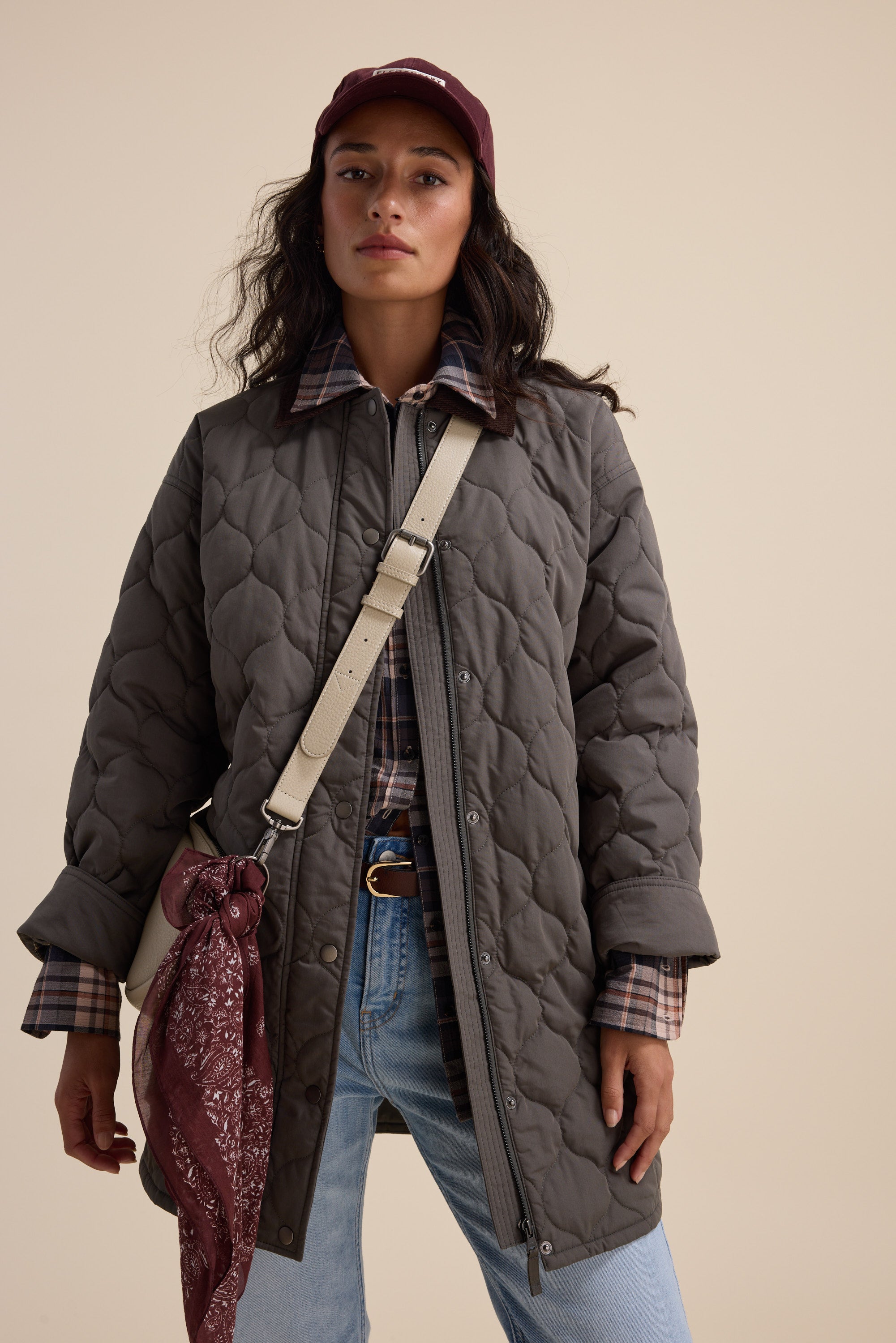 Woman in the Elementary Longline Puffer, plaid shirt, jeans, cap, and crossbody bag.