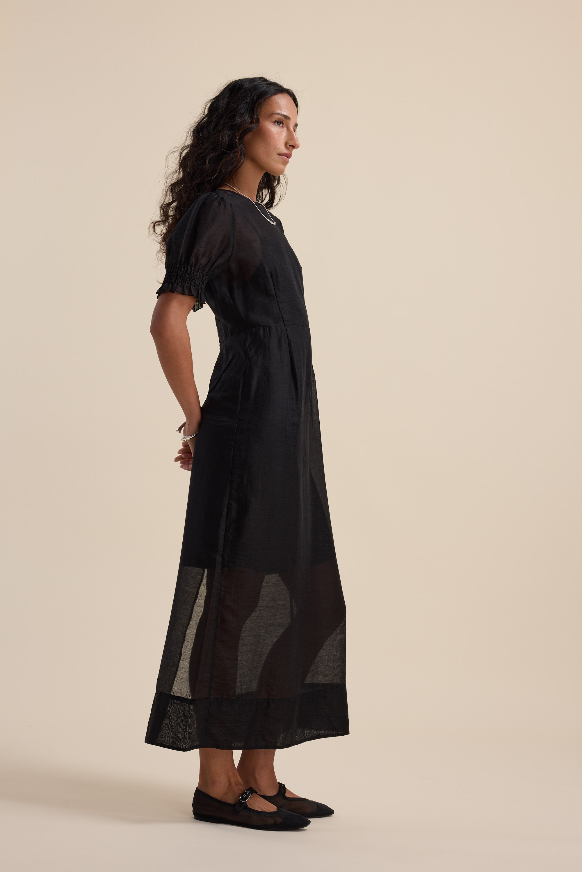 Model in Max’s Sheer Layering Dress, black, standing sideways against a beige background.