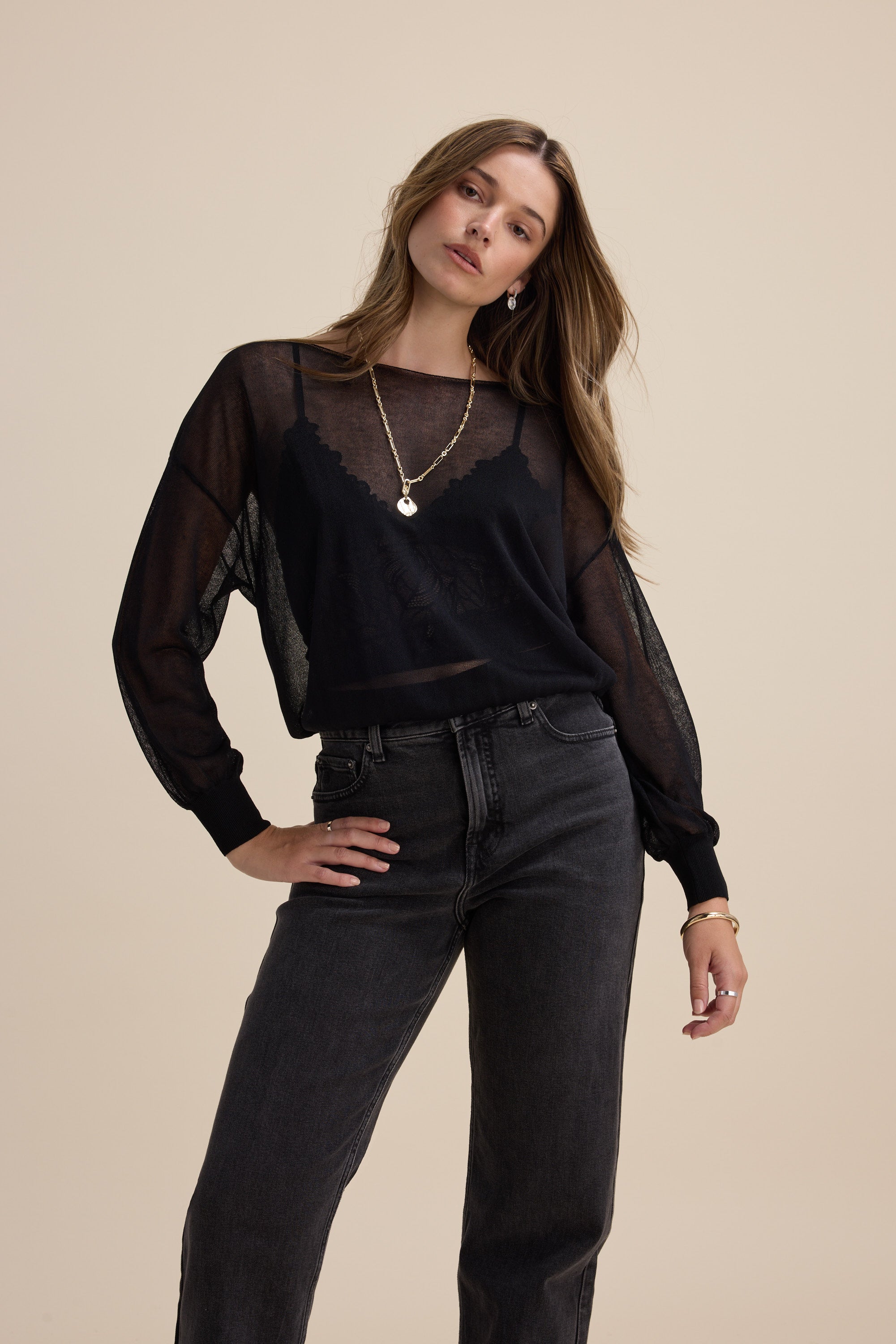 Woman wears Max Addie Sheer Knitted Top with high-waisted dark jeans against beige background.