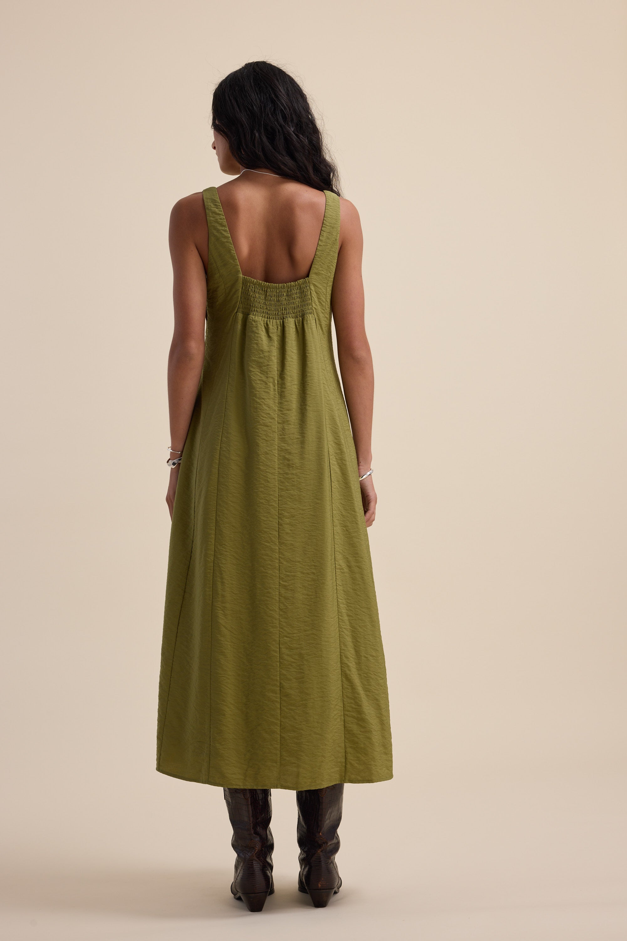 Woman in a Nina Fit and Flare Dress by Max, dill green, sleeveless, with brown boots, rear view.